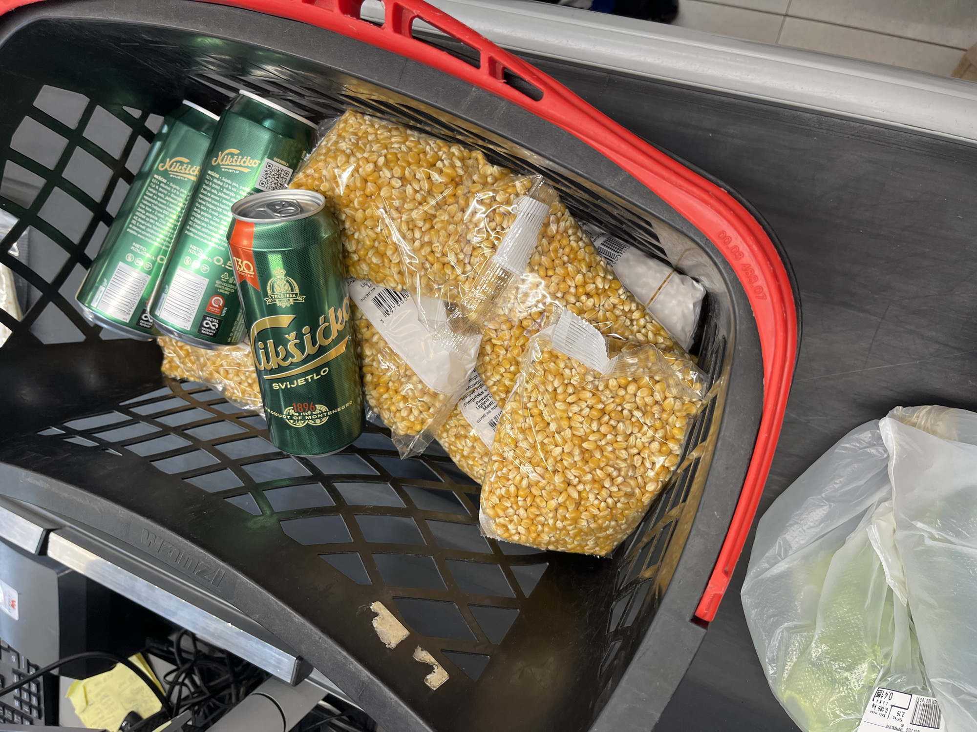 A black shopping basket with a red handle, manufactured by "Wanzl," is depicted resting on a dark grey surface, likely a supermarket checkout counter or conveyor belt. Inside the basket are several items. Three green cans of "Nikšićko SVIJETLO" beer are prominently featured, clearly stating "PRODUCT OF MONTENEGRO," which aligns with the given location context of Budva. Adjacent to the beer cans are multiple clear plastic bags filled with unpopped yellow popcorn kernels. A white plastic bag containing what appears to be green produce is partially visible on the right side of the basket.
The scene suggests a shopping event, with groceries ready for purchase at a retail store. A sticker on the white plastic bag displays "CENA 2,19" (price 2.19) and "0,186 kg" for an item. The red handle of the basket has the code "00.03 488.07" embossed on it. No people, specific time of day, weather conditions, or emotions are visible in the image.
Visible text includes:
- On beer cans: "Nikšićko," "SVIJETLO," "TREBJESA," "1896," "PRODUCT OF MONTENEGRO," "SKENIRAJ," "NETO KOLIČINA," "LIMENKE," "METALL."
- On popcorn bags: Partially visible text such as "Hranjive vrijednosti" (nutritional values), "Energetska v" (energy value), and "cuvati na suvom" (store in a dry place).
- On the white plastic bag sticker: "0,41", "CENA 2,19", "0,186 kg".