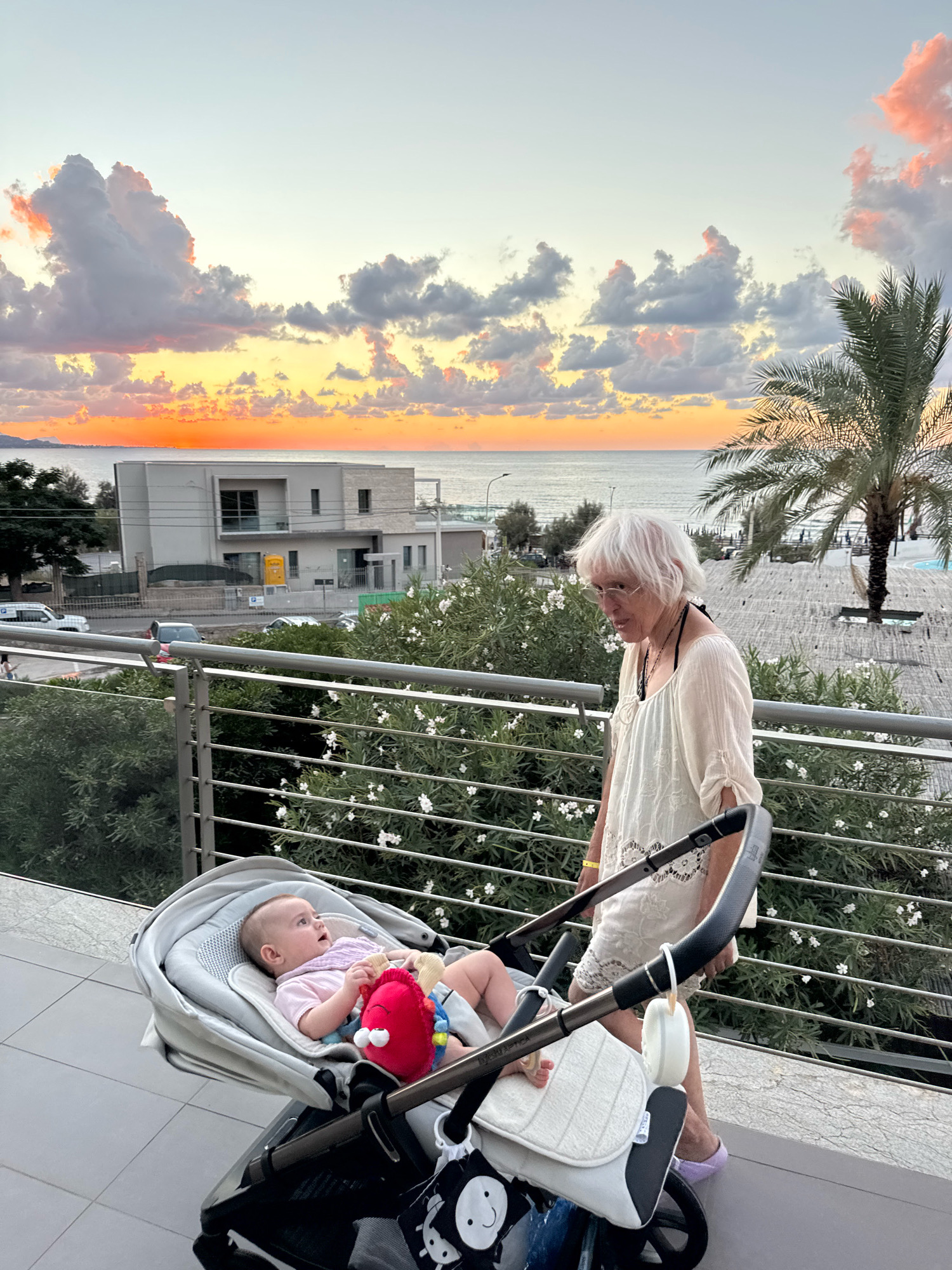 The image depicts a sunset scene on a balcony overlooking the sea in Isola delle Femmine, Italy. A woman with white hair, wearing glasses and a light-colored top, stands near a baby in a stroller. The baby is lying in the stroller, facing upwards, and appears to be awake. The woman is looking down and to her left, while the baby is looking up and to its right. In the background, there is a modern building, trees, a palm tree, and the sea. The sky is filled with colorful clouds, indicating a sunset. The balcony has metal railings and a tiled floor.