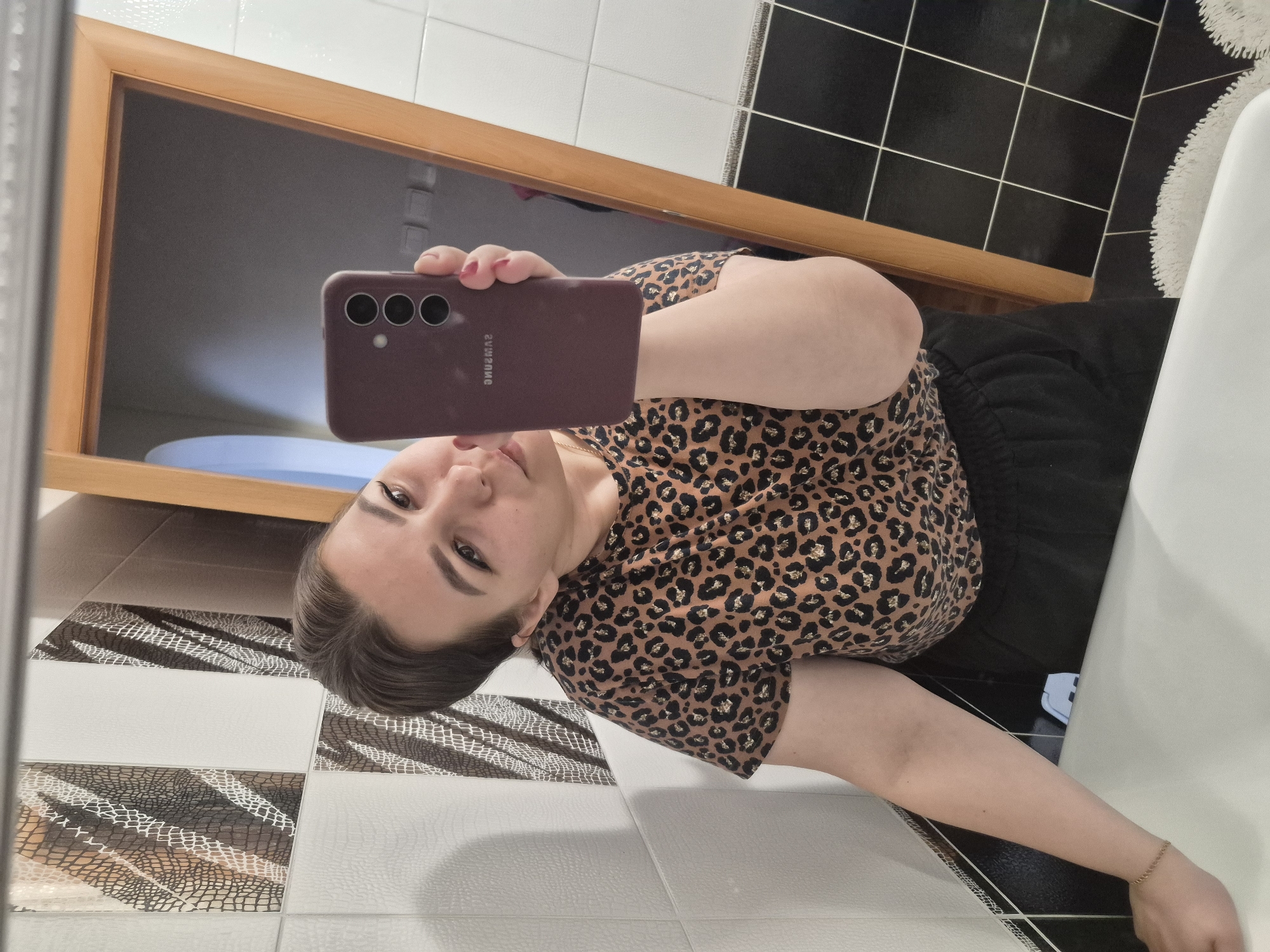 A woman is taking a selfie in a bathroom. The bathroom has a mirror with a wooden frame, black and white checkered tiles on the wall and floor, and a white sink. The woman is wearing a brown and black leopard print shirt and black pants. She is holding a maroon Samsung smartphone in her right hand. The reflection in the mirror shows part of a blue toilet seat. The city of Tobolsk, Russia is not present in the image.