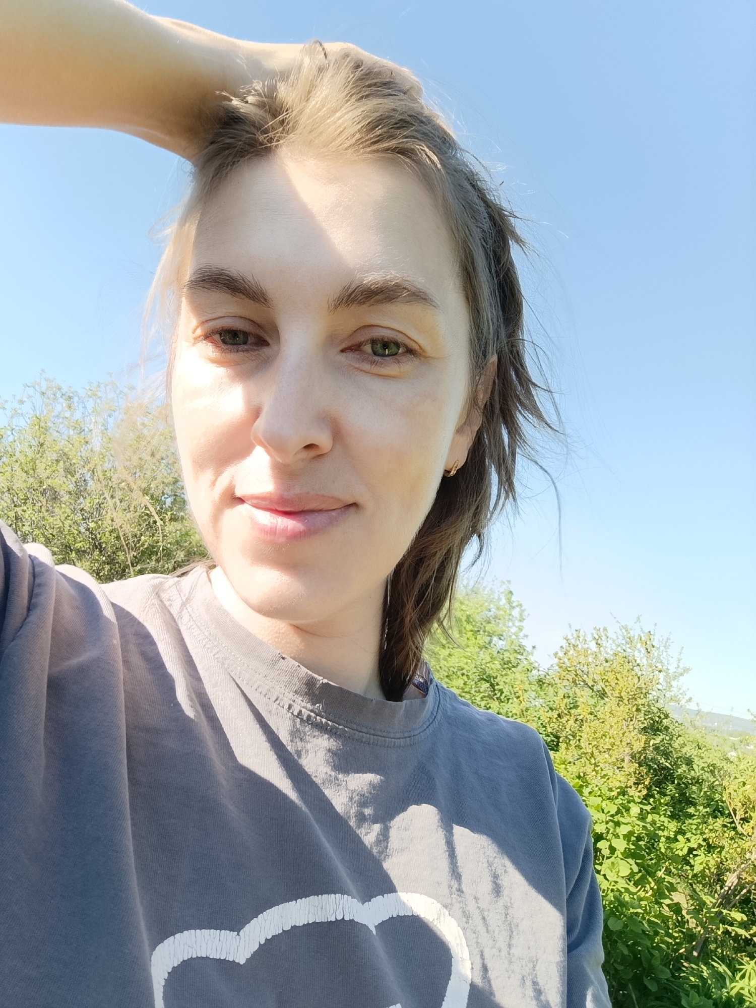 A selfie was taken outdoors on a sunny day in a location with greenery and a clear, blue sky. The focus is on a young woman who is fair-skinned with light brown or blonde hair pulled back, she is wearing a grey t-shirt with a white print. Her left arm is raised, with her hand positioned on her head. Green trees and bushes are visible in the background. The location is not explicitly mentioned in the image, so providing a precise location in Chaikovsky, Russia, is not possible based solely on the image content.