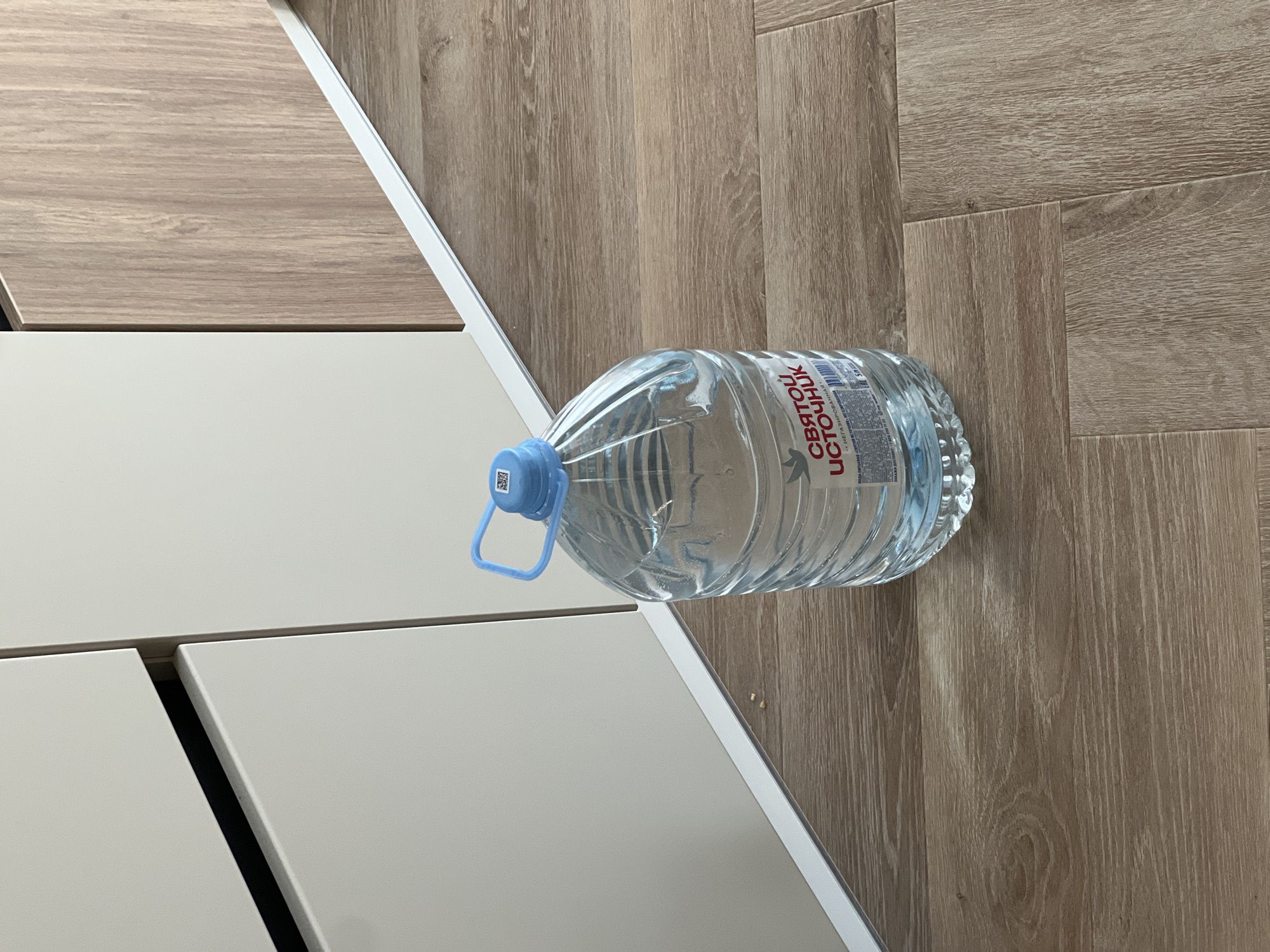 The image shows a 5-liter bottle of "Святой Источник" (Holy Spring) drinking water on a wooden floor. The bottle has a blue cap and handle. Part of a white and light brown cabinet or furniture piece is visible on the left side of the image. The scene is indoors. The location is Новое, Russia based on the brand of the water bottle.