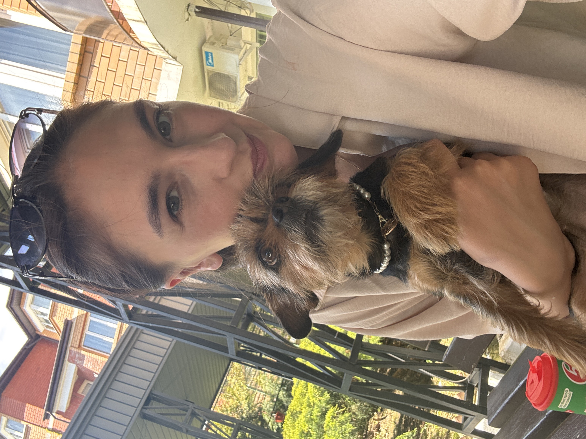 A light-skinned woman with brown hair pulled back is holding a small, terrier-type dog with brown and black fur. The dog is wearing a black collar with white pearls and a small charm. The woman is smiling and looking directly at the camera. Sunglasses are perched on her head.
The background shows an outdoor setting, possibly a patio or veranda, with a metal framework overhead. In the distance, there is a red brick building with windows and greenery suggesting a garden or park. The lighting indicates it is daytime.
Based on the visual clues and the user-provided information, the location is likely Sochi, Russia.