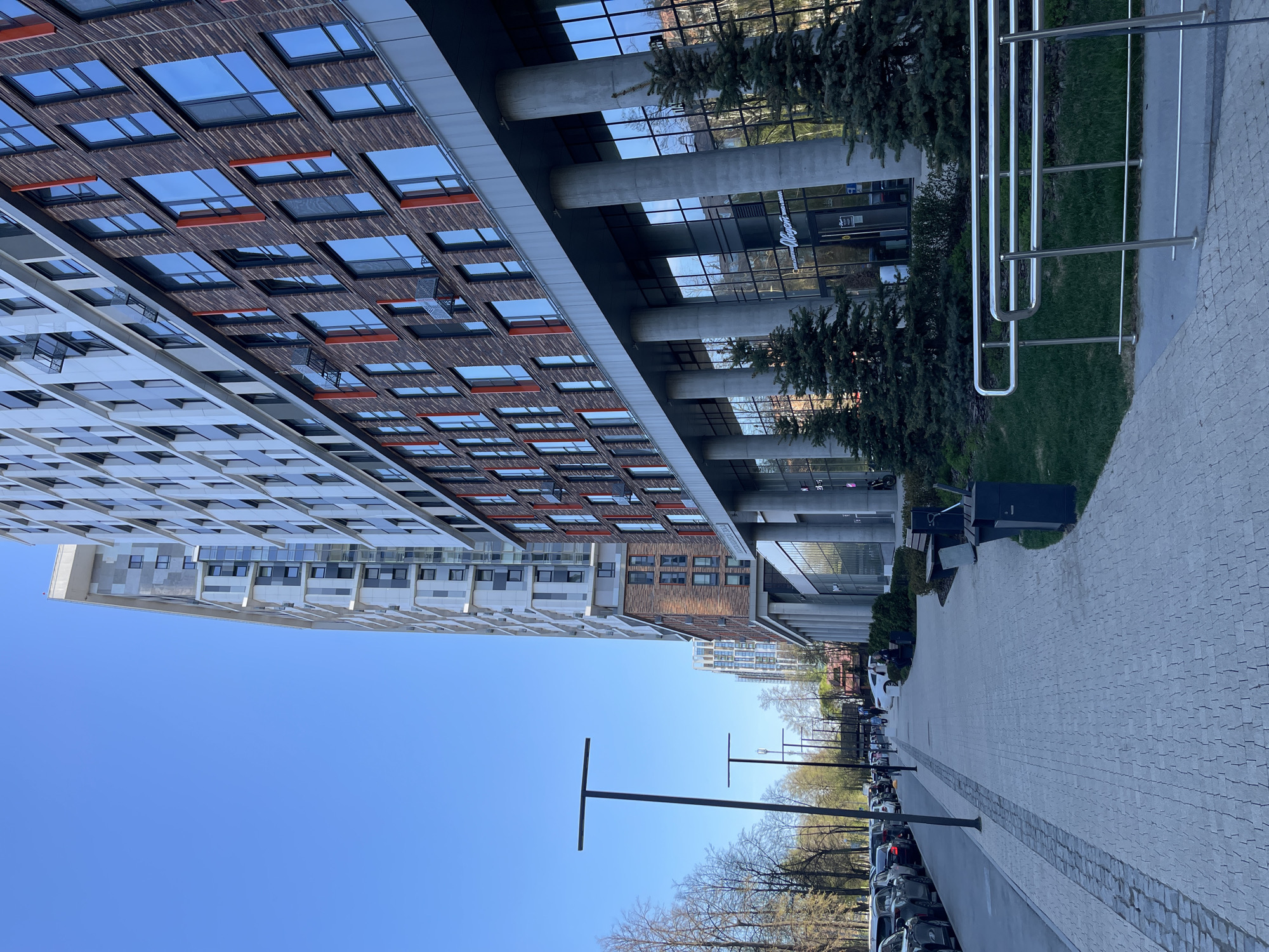 The image depicts an urban scene in Izhevsk, Russia. Two modern buildings stand adjacent to each other; one is characterized by a white facade with recessed windows, while the other features a reddish-brown brick-like exterior with windows reflecting the sky. A glass-enclosed walkway supported by concrete pillars connects the buildings.
A paved walkway runs alongside the buildings, lined with trees and lamp posts. Parked cars are visible along the edge of the walkway. On the opposite side of the walkway from the street, some metal railings. The sky is clear and blue.