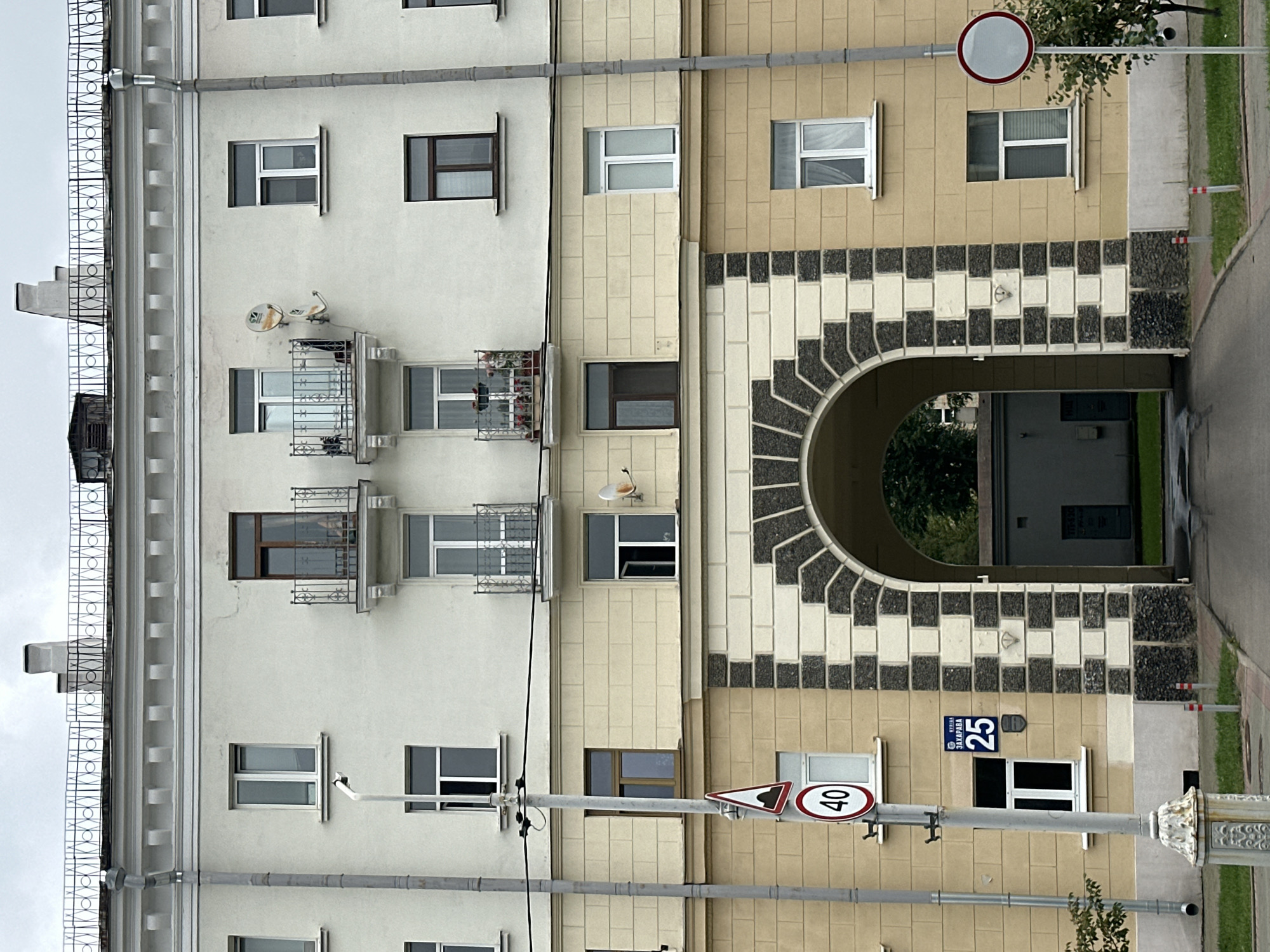 The image shows the facade of a multi-story building in Minsk, Belarus, with an arched entryway in the center. The building has a light cream-colored upper section and a beige lower section. Several windows are visible on both sections, some with balconies on the upper level. A street sign indicating "40" for speed limit and a road hazard sign are mounted on a pole to the left of the entryway. The address plaque on the building reads "вул. Захарава 25" which translates to "Zakharova Street 25". The entryway leads to a dark alleyway with a building visible in the distance. The sky is overcast.