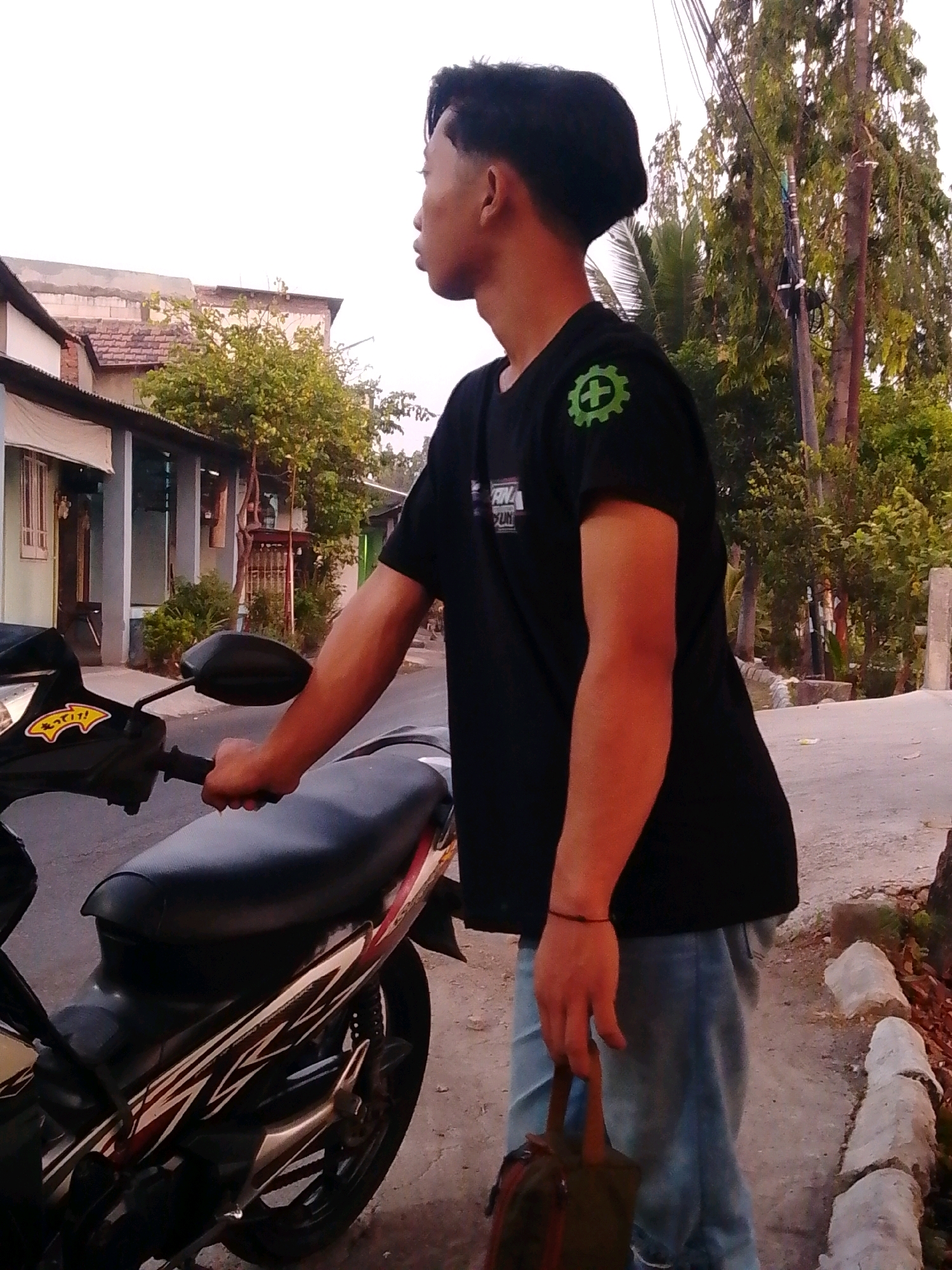 A young man with dark hair, wearing a black t-shirt with a green gear emblem and light blue jeans, stands next to a black motorcycle in Tambak Sumur, Indonesia. He is holding a brown bag in his right hand and resting his left hand on the motorcycle's handlebar. The background shows a street with buildings and trees, typical of a residential area. The lighting suggests it is daytime, possibly late afternoon due to the warm tones. The motorcycle's seat is black with red and gold decals on its side panel. The man is looking to his right, away from the camera.