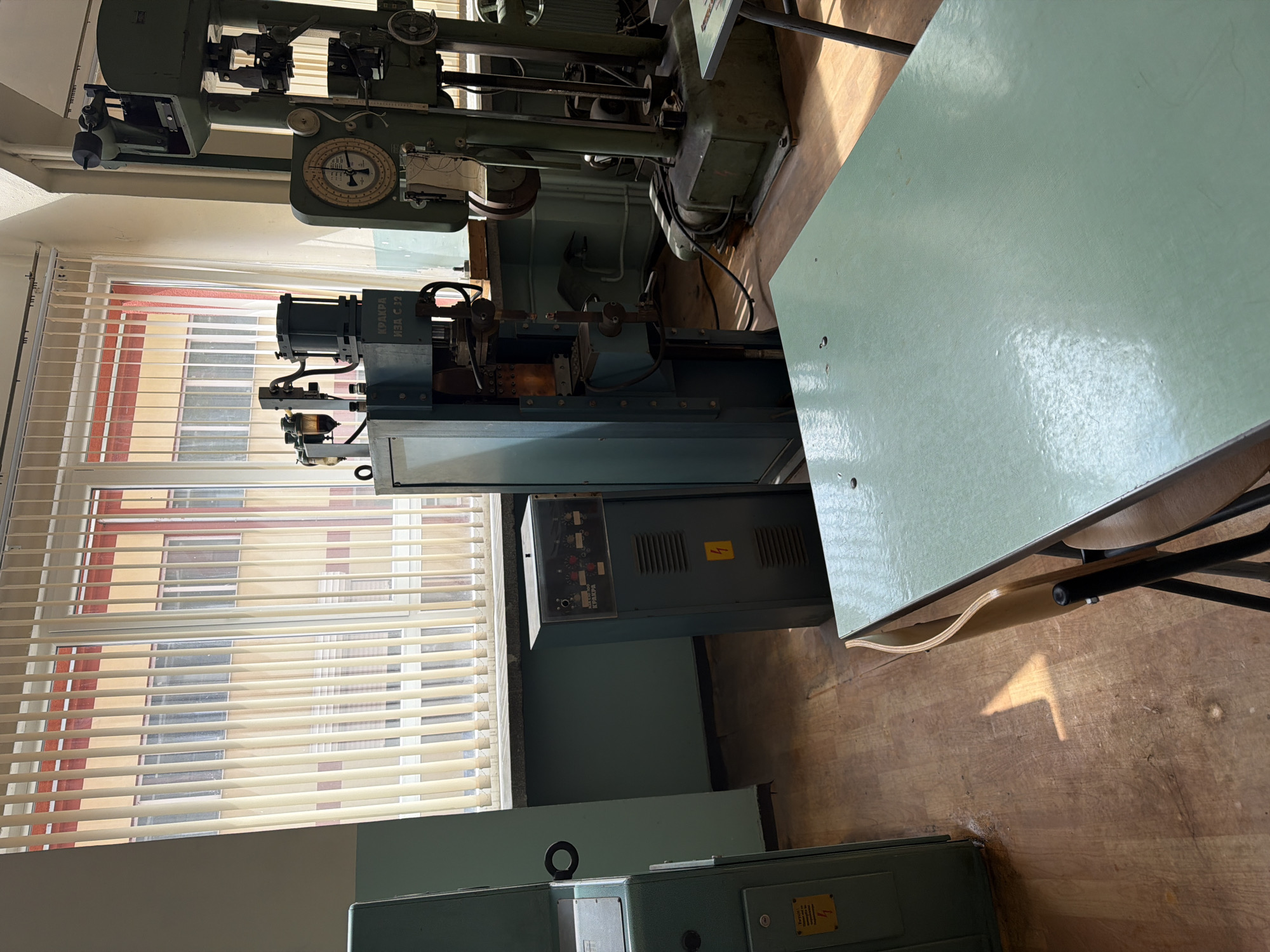 The image shows a room with a large, green, industrial machine next to a window with closed blinds. The machine is likely a lathe or similar metalworking tool, given its construction and dials. A green table is next to the machine. The floor is wooden. "KRAKRA" is visible on the machine indicating that it was manufactured in Bulgaria. Considering the vintage style of the machine, the location might be a vocational school or old factory in Sofia, Bulgaria, given its history as a center for industrial production.