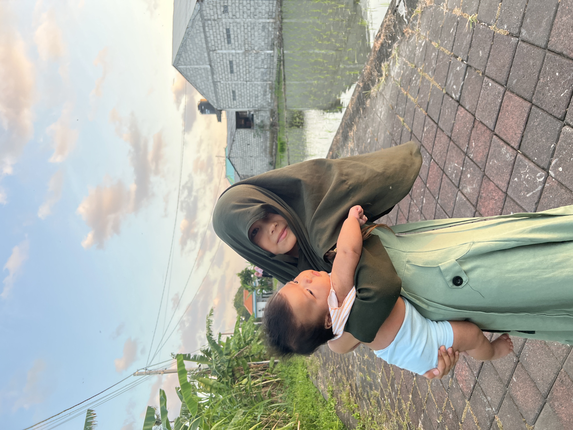 The image captures a young girl or teenager holding an infant in a rural or semi-rural setting in Mengwi, Indonesia, during the golden hour. The girl, positioned in the center-right, wears a long-sleeved light green dress featuring a dark button detail and a dark green hijab that covers her head and upper torso. She looks directly at the camera with a slight smile. The infant, appearing to be several months old, wears a white and orange striped top and light blue shorts, and is looking towards the viewer's left. The girl cradles the infant, supporting its back and legs.
The foreground displays a paved pathway constructed from rectangular, interlocking blocks of reddish-brown and dark grey, exhibiting various white marks and sparse vegetation. To the left of the path, dense green tropical foliage, including prominent banana plants, is visible, along with a wooden utility pole supporting multiple overhead wires. In the midground to the right, a flooded rice paddy field with numerous young green rice shoots emerges from the water, reflecting the sky. The background reveals several block-built structures with simple roofs, including a larger grey concrete block building to the far right. The sky overhead is a light blue with scattered cumulus clouds, subtly tinged with soft orange and pink hues, characteristic of late afternoon or early evening light.