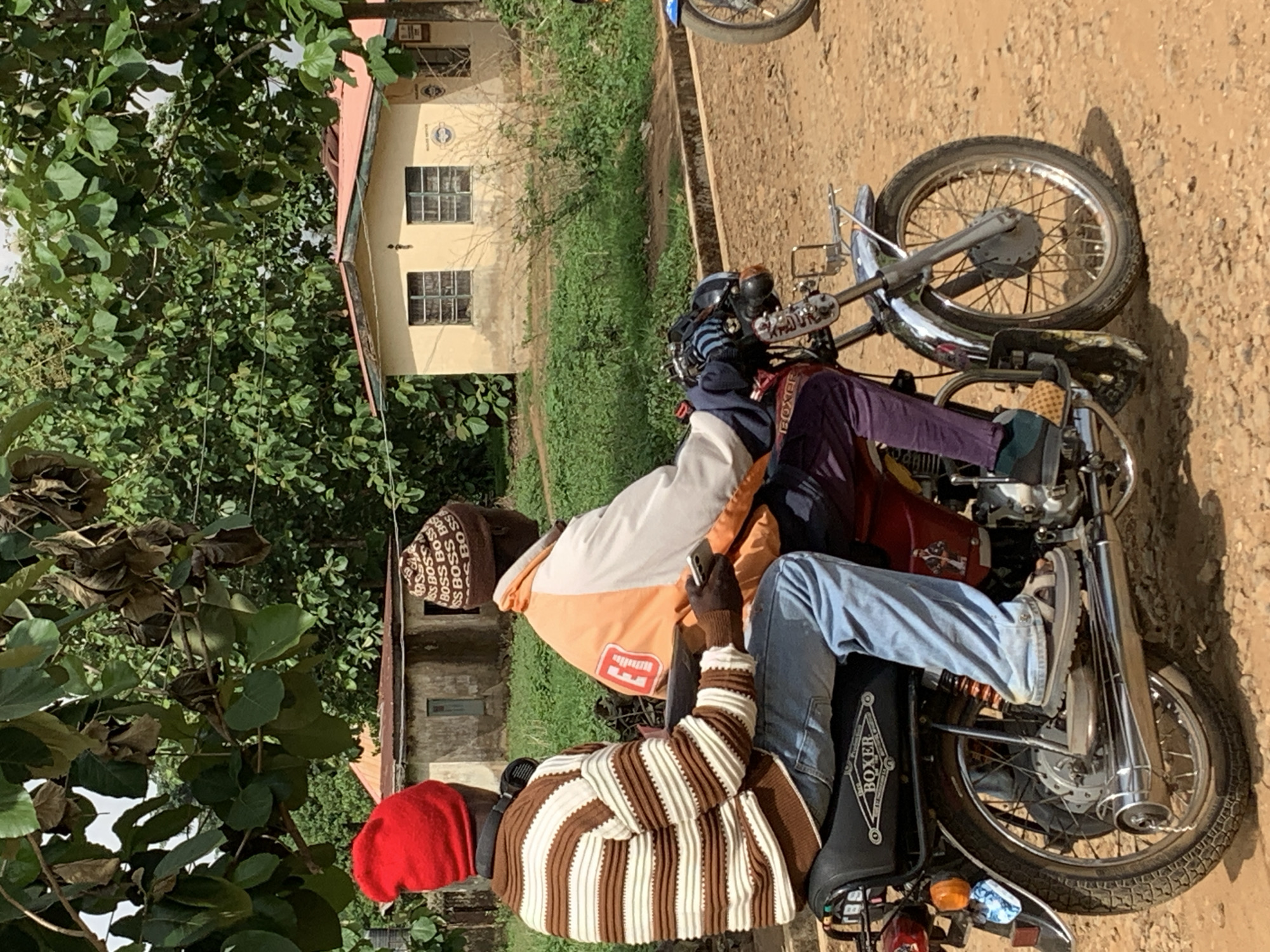 The image shows two motorcycles parked on a dirt road in Maigizo, Nigeria. One motorcycle is a red model with the "BOXER" logo visible. Two men are sitting on the motorcycles. One man is wearing a red cap and a brown and white striped sweater. The other man is wearing a brown "BOSS" patterned hat and an orange and white jacket. One man is holding a cell phone. A tan building with two visible windows is in the background. Trees and green vegetation surround the building.