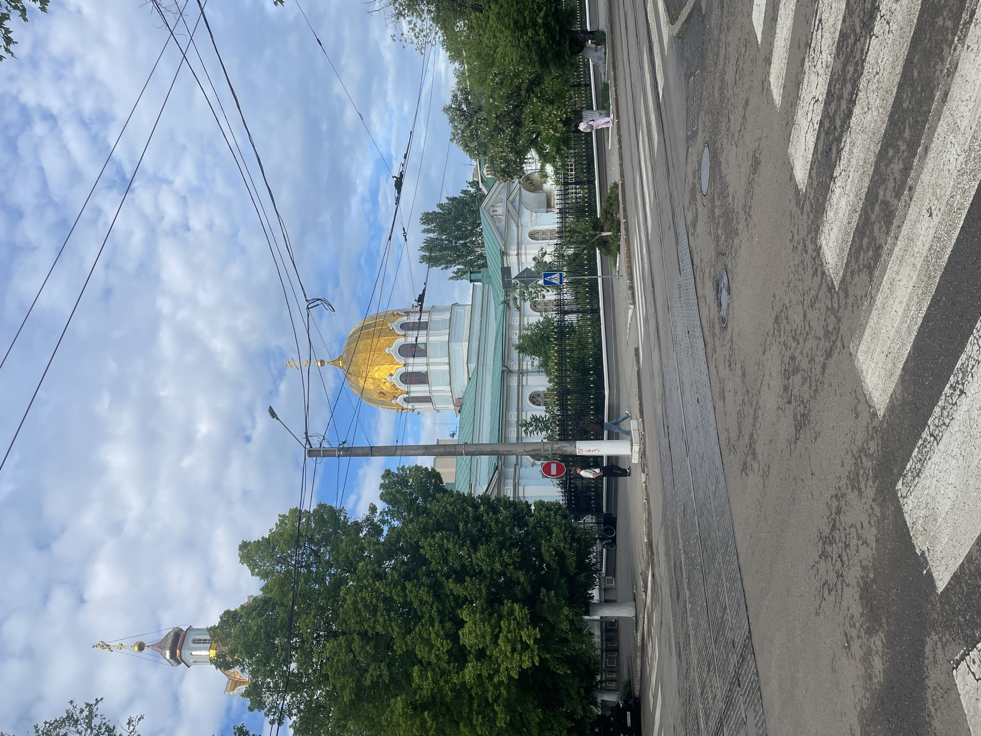 The image shows a street in Mykolaiv, Ukraine. A large Orthodox church with a prominent gold dome is visible on the left. Power lines crisscross the sky above. Trees line the street. On the right is a zebra crossing over a wet street with tram tracks. Two people can be seen near the church fence. One person is standing and the other is sitting. There are also parked bicycles nearby. The sky is cloudy with patches of blue.