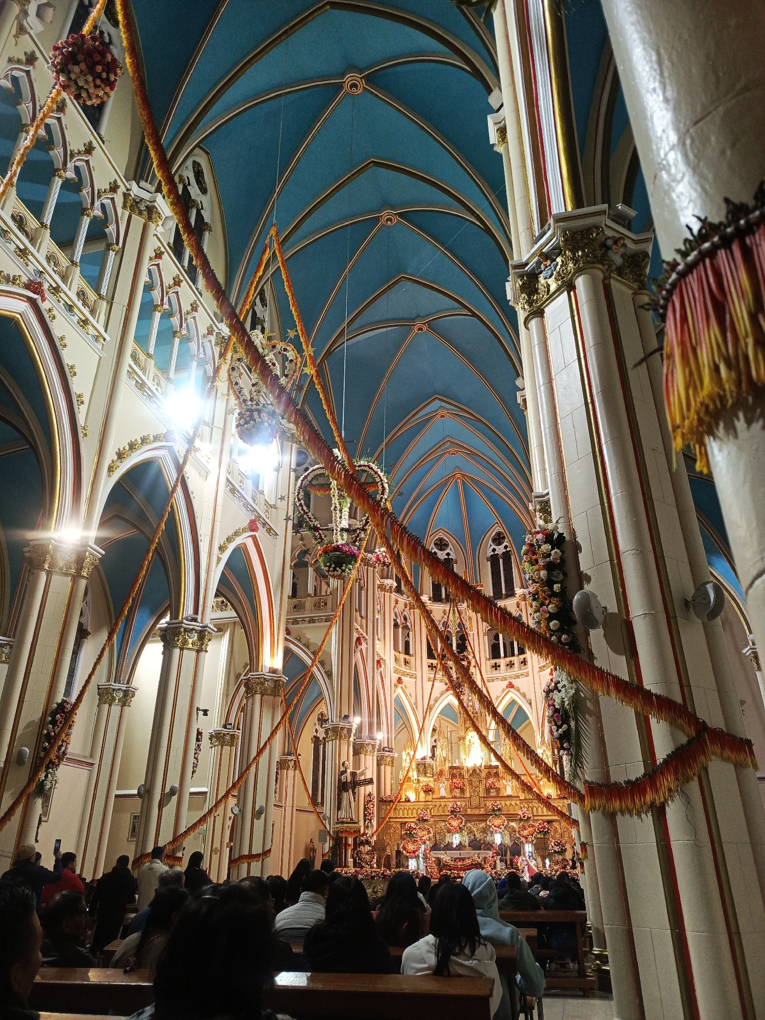 The image depicts the interior of a church filled with people seated in pews. The church has a high, vaulted ceiling painted blue, with ornate architectural details and columns decorated with gold accents. Garlands of orange and yellow decorations hang across the space. The altar is adorned with flowers and religious figures. The location is the church in Unknown, Unknown.