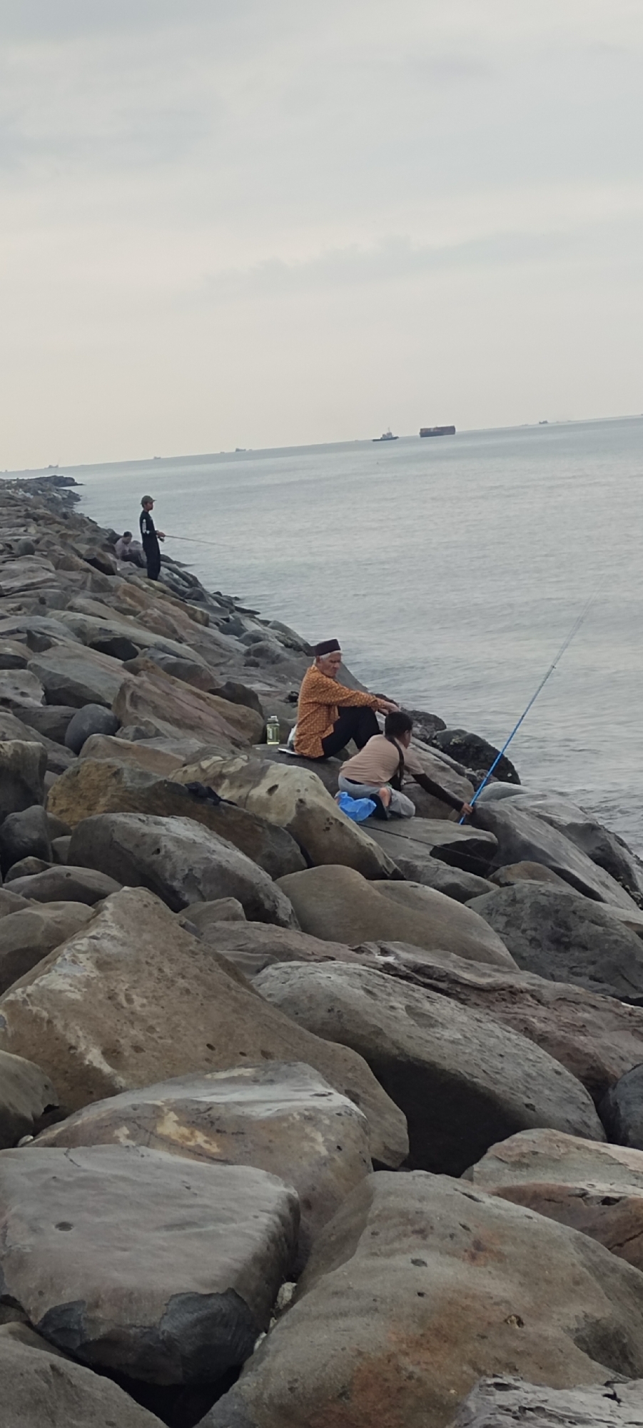 A group of people are seen fishing on a rocky breakwater. The water is calm, with several ships visible in the distance. The sky is overcast. The scene is located at the Lhokseumawe City breakwater in Indonesia. One person, an older man with a traditional cap, is seated and fishing. Another younger person is kneeling and also fishing. A third person stands further down the breakwater, holding a fishing rod. The breakwater is constructed from large, grey rocks.