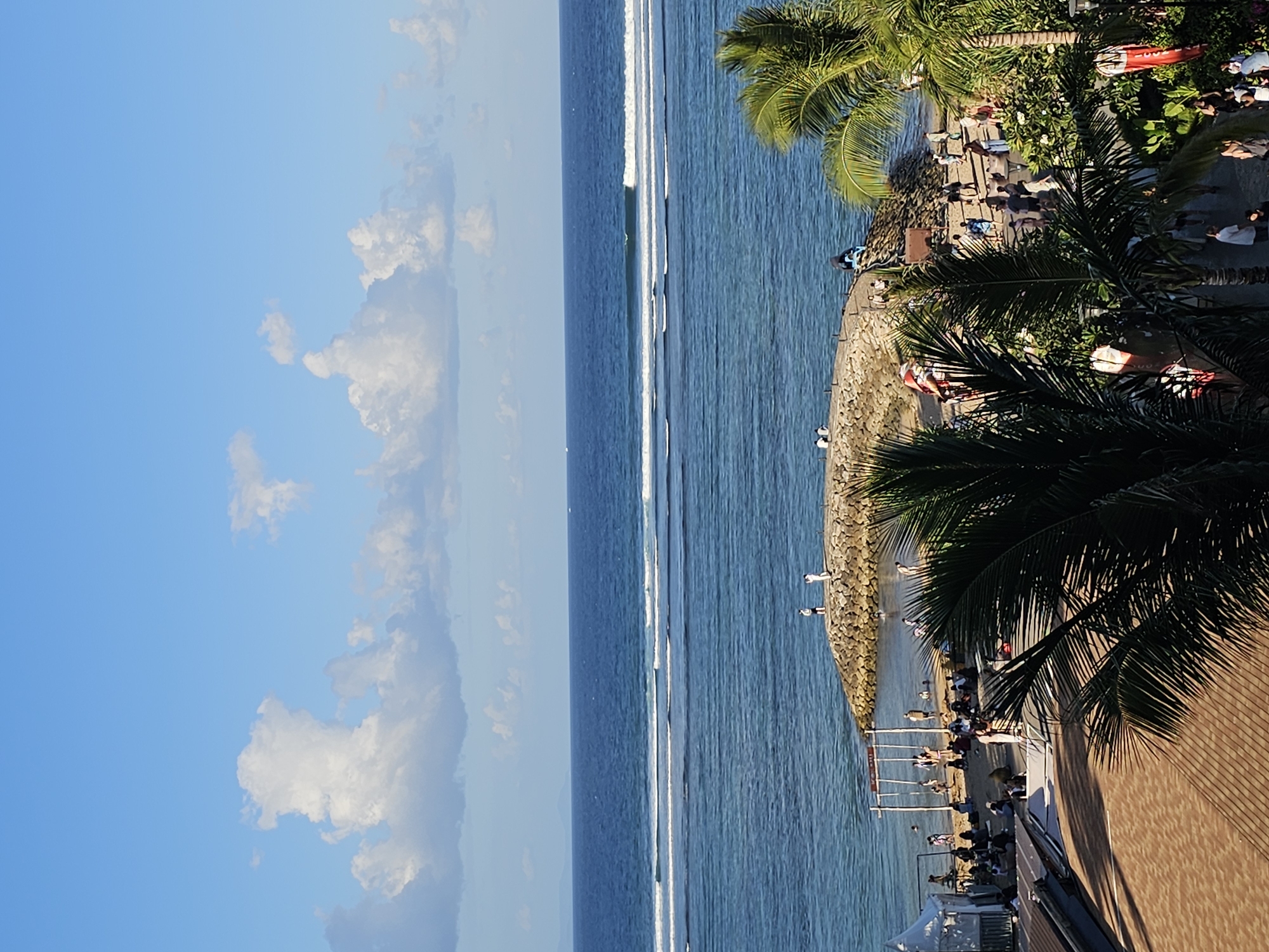 This is a high-angle, full-frame shot of a tropical beach scene, likely in the Denpasar area of Indonesia, depicting a sunny day with a clear blue sky dotted with white cumulus clouds. The ocean occupies the upper two-thirds of the image, with distinct sets of white waves breaking at different distances from the shore, indicating a shallow reef.
The foreground and lower third of the image are dominated by palm trees with lush green fronds, framing a bustling beach area and a small jetty. The beach itself is sandy, with numerous people scattered across it, some sitting, some standing, and some walking. A rocky jetty extends into the water, with several individuals visible on it, some observing the ocean and others walking.
On the left side of the beach, there appears to be a small structure, possibly a tent or shelter, and a swing set-like structure made of poles. Further back on the beach, a crowd of people is gathered. The lower right corner shows more palm trees and foliage, and a paved pathway with more people walking. The overall scene suggests a popular tourist destination.