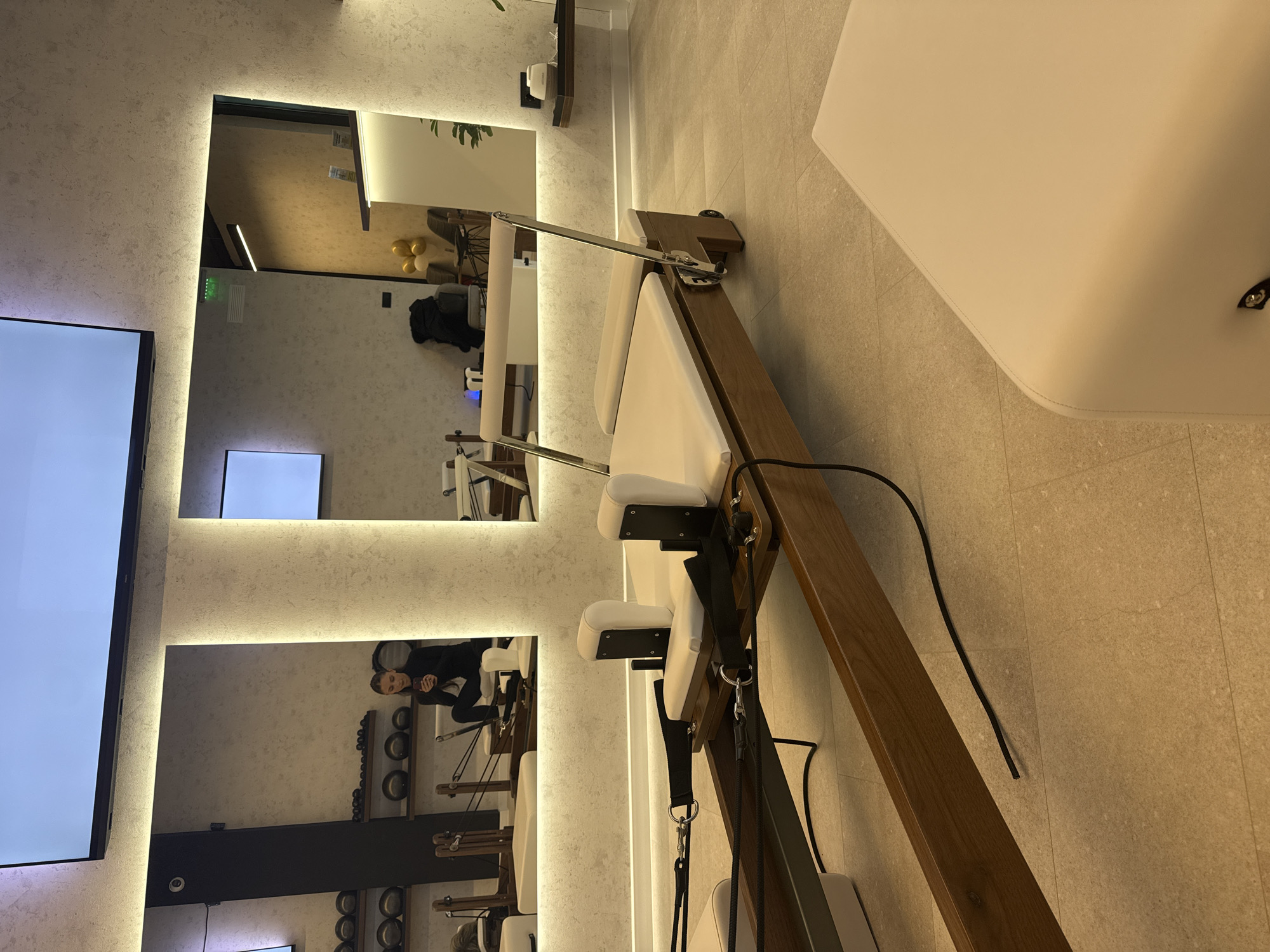 The image shows the interior of a Pilates studio, as evident by the Pilates equipment, including a wooden reformer with a white cushion and adjustable parts. A person, seemingly a woman, is visible in the reflection of one of the mirrors, possibly taking a selfie. The studio has a neutral color palette with tile flooring and textured walls, highlighted by vertically aligned recessed lighting. Multiple mirrors of varying sizes are strategically placed, reflecting different parts of the room. A television screen and shelving with weighted balls are visible. The location is suggested to be Sofia, Bulgaria.