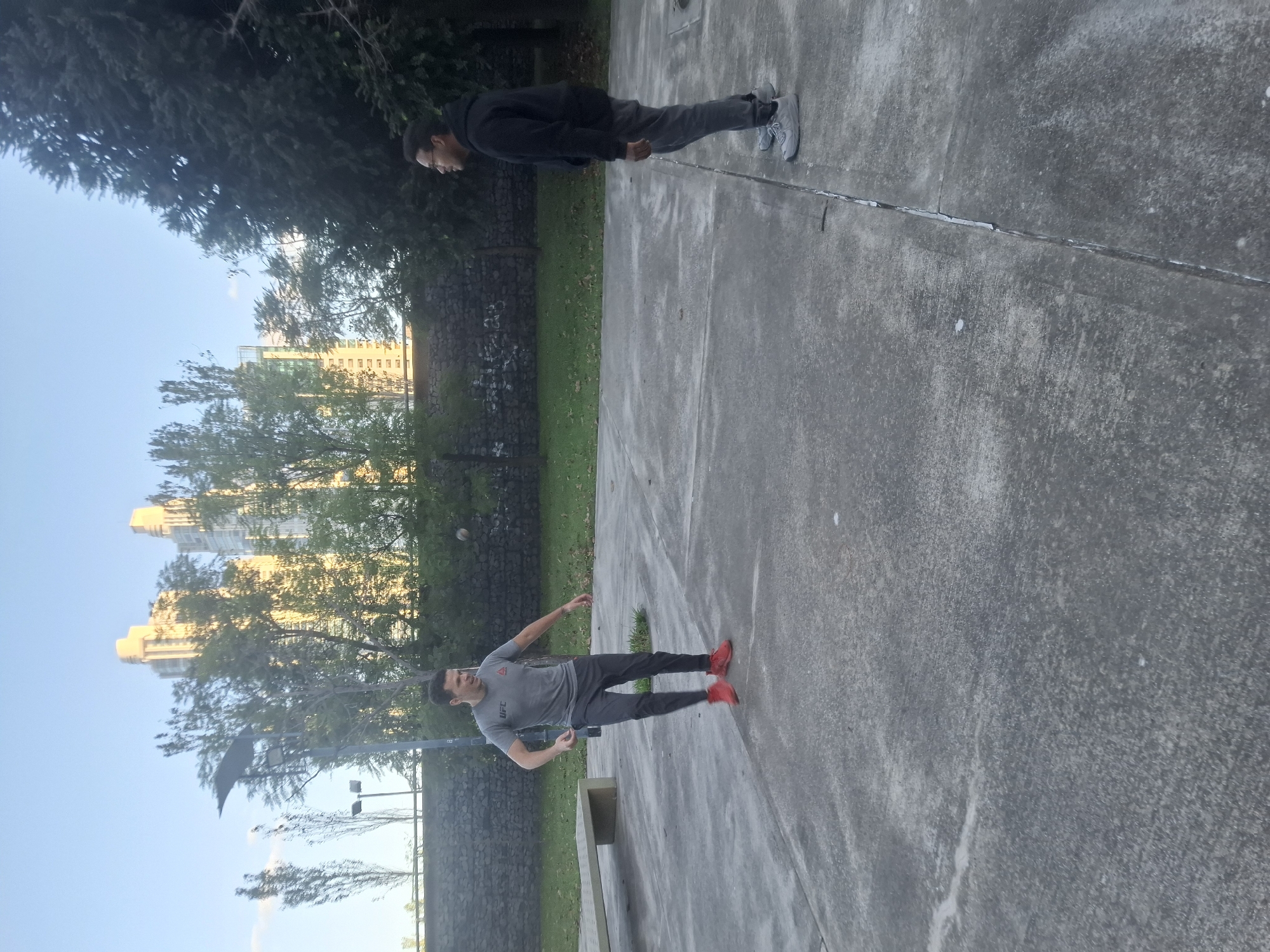 Two men are outdoors in a public space with a paved surface. One man is wearing a dark hoodie, dark pants, and light-colored shoes. The other man is wearing a grey t-shirt with a logo, dark pants, and red shoes. There is a pole or rod-like object lying on the ground between them. The background includes trees, buildings, and a stone wall. It is daytime. The location appears to be in the city of Buenos Aires, Argentina, judging by the style of architecture of the buildings in the background.
