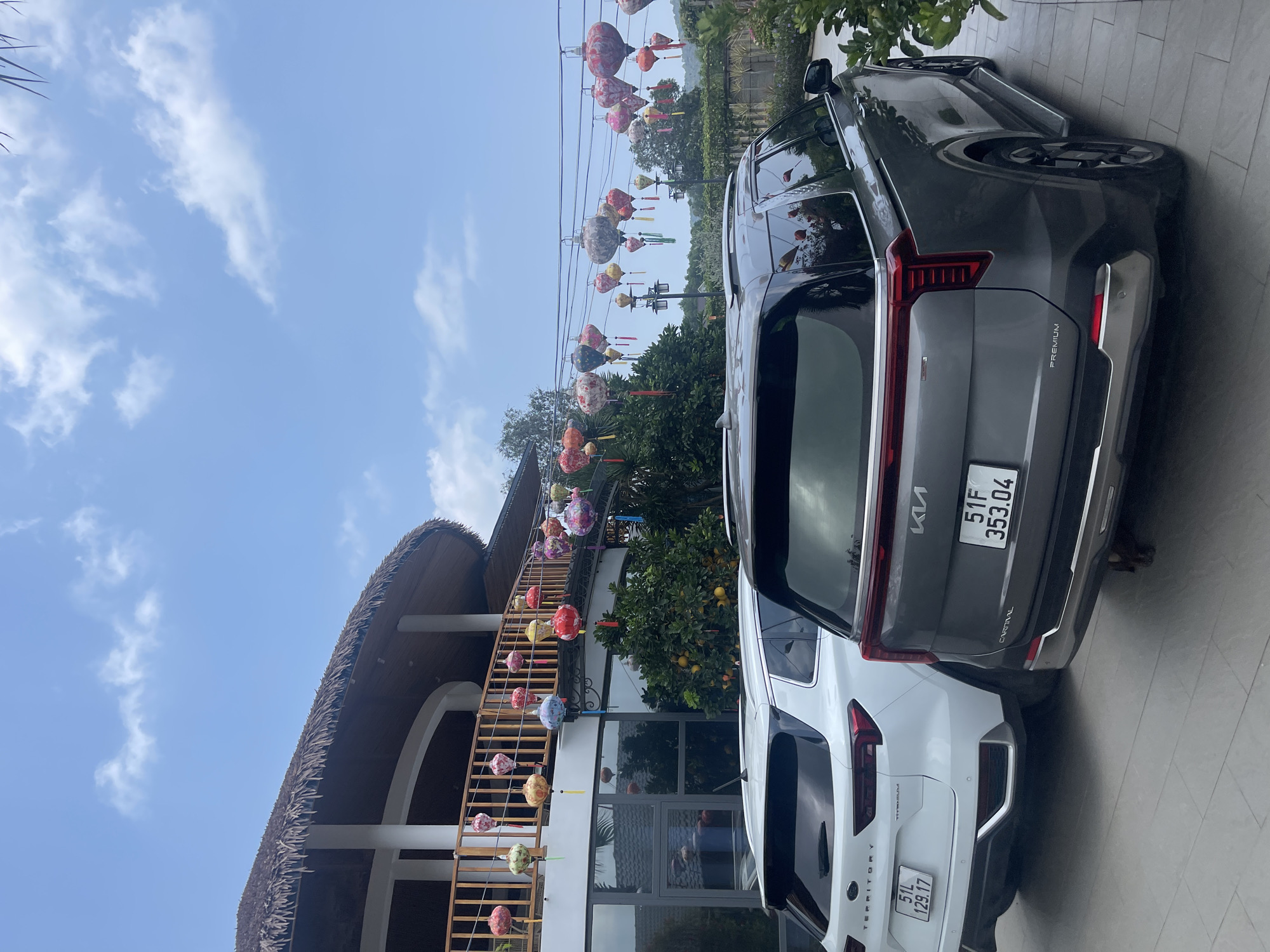 The image shows the exterior of a building in Phú Quốc, Vietnam, under a partly cloudy blue sky. The building has a distinctive thatched roof and a wooden staircase decorated with lanterns. Two parked cars, a white Ford Territory and a gray Kia Carnival, are positioned closely together. The license plate of the Kia Carnival is visible and reads "51F 353.04", while the Ford Territory's license plate reads "51L 129.17". Overhead, a series of lanterns are strung between power lines. An orange tree is situated near the building. The scene appears to be a bright, sunny day.