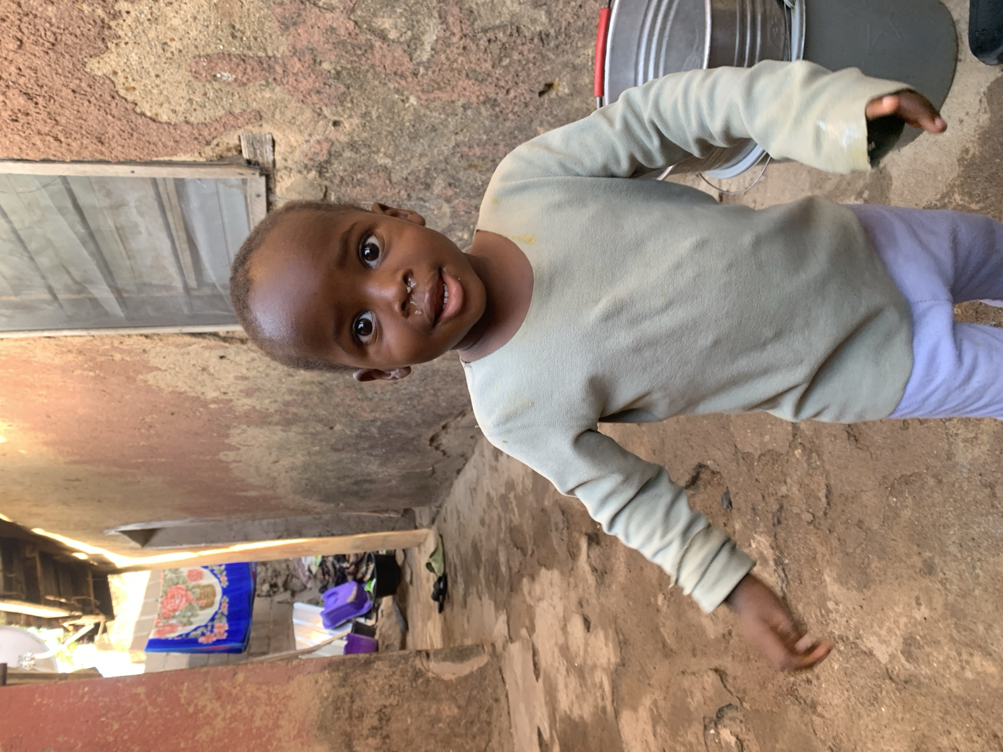 A young child, likely between 2 and 4 years old, stands on a rough, cracked, and uneven concrete surface within a building or structure. The child has brown skin, short hair, and appears to be in good health, despite some nasal discharge. The child is wearing a light-colored, long-sleeved shirt and light purple pants. The background features aged walls, suggesting an older or possibly run-down building. Visible in the periphery are items like metal buckets, laundry (a blue sheet with a floral pattern), and a purple bag. The location is Jos, Nigeria.