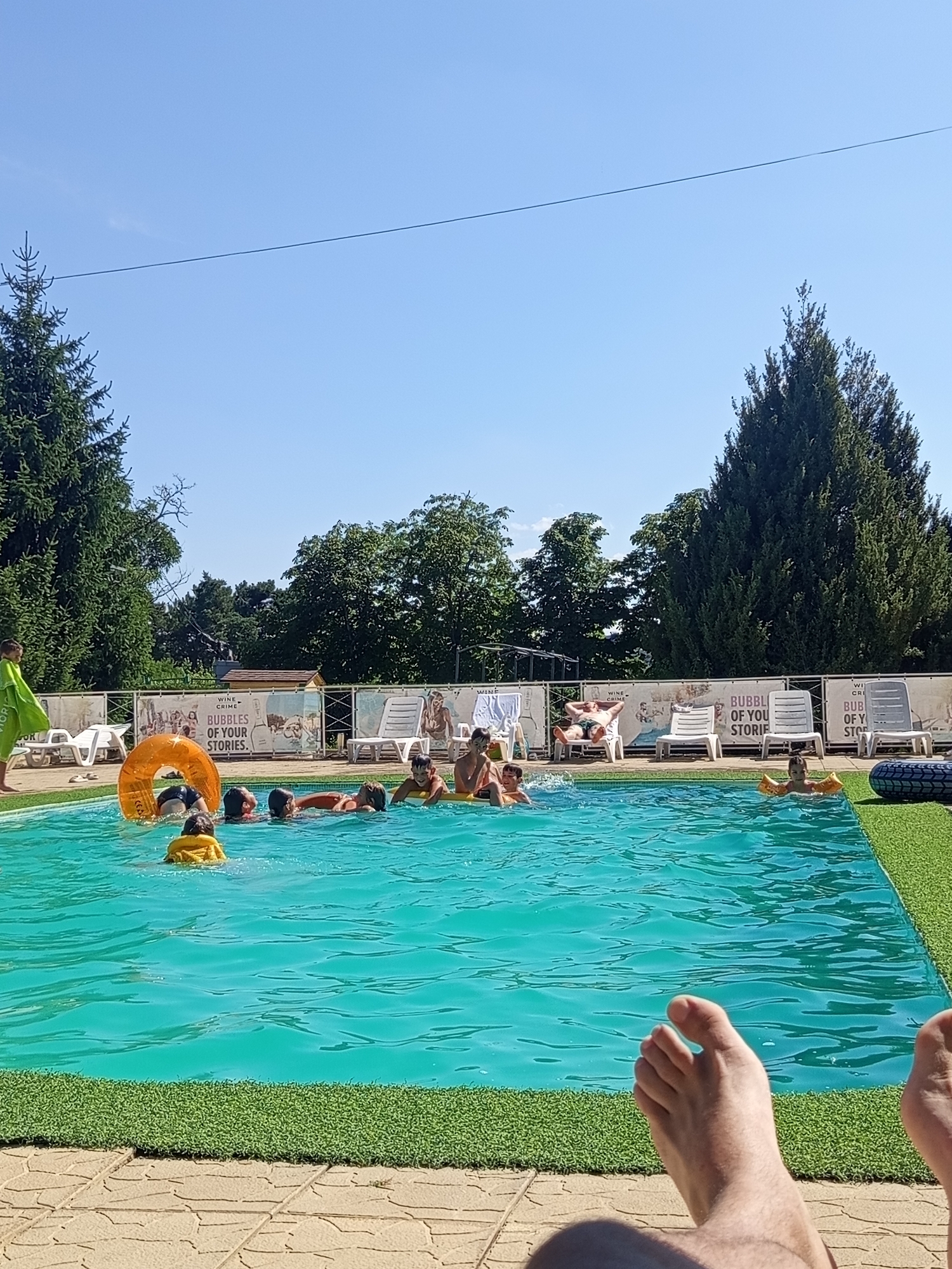 The image depicts an outdoor swimming pool on a sunny day, likely in Molovata Nouă, Moldova, given the context. Several individuals, mostly children and young adults, are visible in the bright turquoise water. Some are swimming, while others are interacting with inflatable toys, including a large orange ring and yellow armbands. A few adults are relaxing on lounge chairs by the poolside. The pool area is bordered by a textured green surface resembling artificial turf, with a paved walkway made of interlocking stone tiles in the foreground. Lush green trees and a clear blue sky form the background, suggesting a natural, possibly recreational setting. A fence with various banners, one featuring the text "BUBBLE OF YOUR STORIES," separates the pool area from the background. A person's feet are prominently featured in the lower right corner, suggesting a first-person perspective.