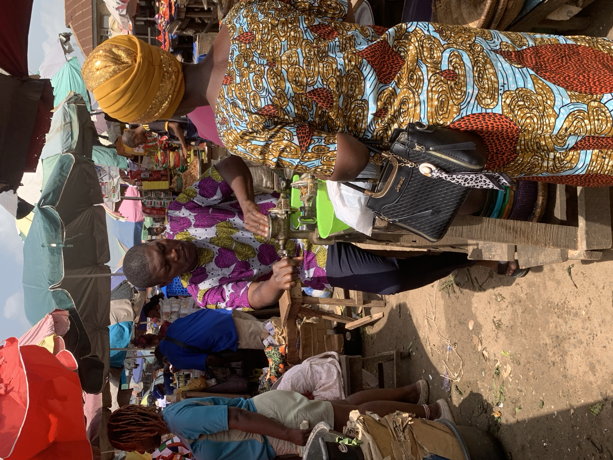 The image shows a bustling market scene in Shagamu, Nigeria. A man wearing a purple and white patterned shirt is operating a piece of machinery, possibly food-related, with a green container nearby. A woman in a colorful patterned dress and a golden head covering is standing beside him, holding a black purse. Market stalls are visible in the background, covered by colorful umbrellas. People are interacting and walking through the crowded market space. The ground appears to be dusty.