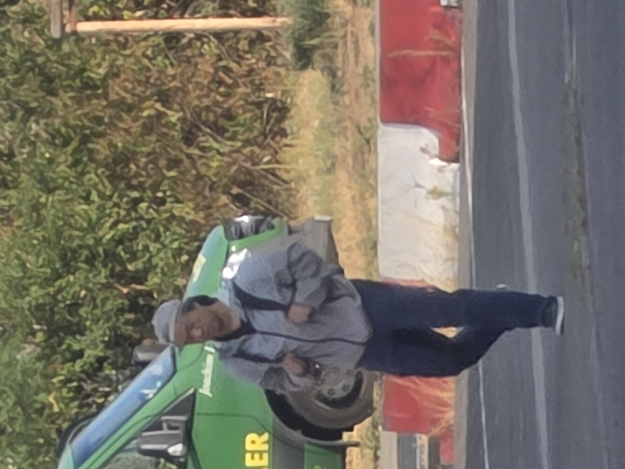 A person is walking across a street in Frankfurt, Germany, in front of a green van and red and white barriers. The person is wearing a gray jacket, dark pants, and a gray cap, and is carrying a backpack. The van has "LER" written on its side in yellow letters and text above it that is partially obscured. The background consists of greenery and a wooden pole.