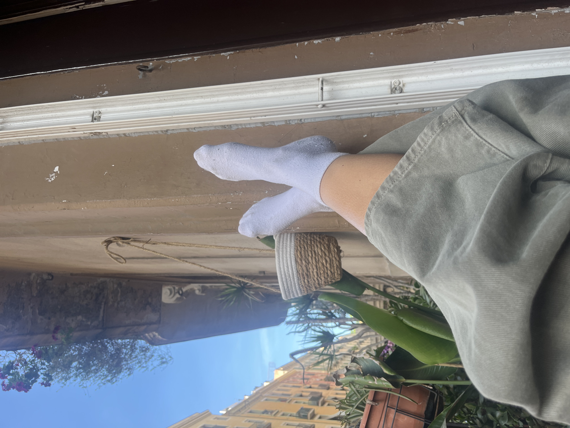 The image shows a person wearing white socks, leaning against a window frame. The person is also wearing green pants. Outside the window, there are plants and a building. The location appears to be Barcelona, Spain.