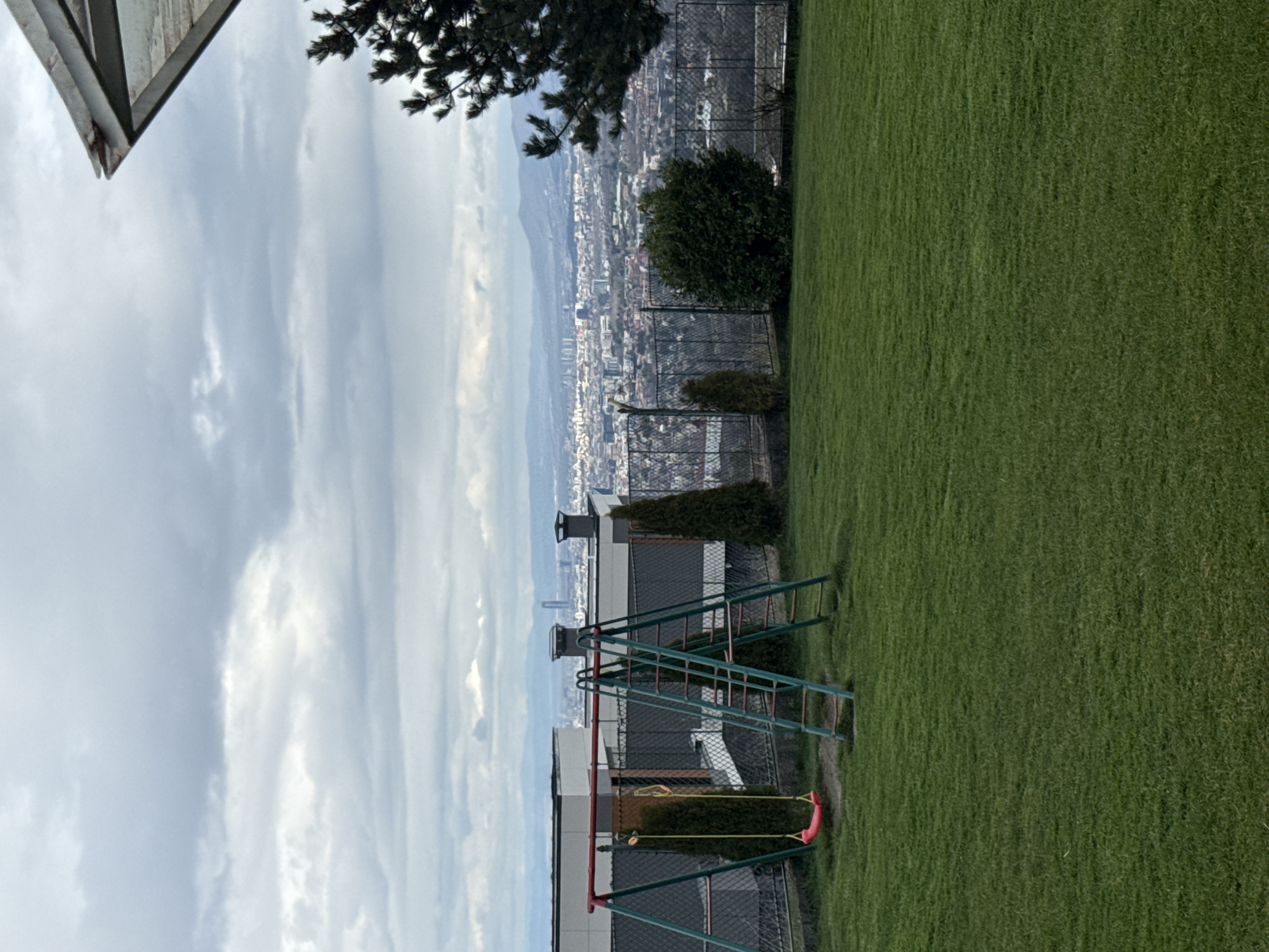 The image depicts a cityscape viewed from an elevated position, possibly a backyard or terrace. In the foreground, there is a green lawn area, bordering a fence. A playground structure with swings and climbing frame is present. Beyond the fence lies a densely populated urban area, blending into the background with a slightly hazy, overcast sky. It is presumed to be in the city of Sofia, Bulgaria, based on the urban setting and mountainous backdrop consistent with Bulgaria's capital. No people are visible in the frame.