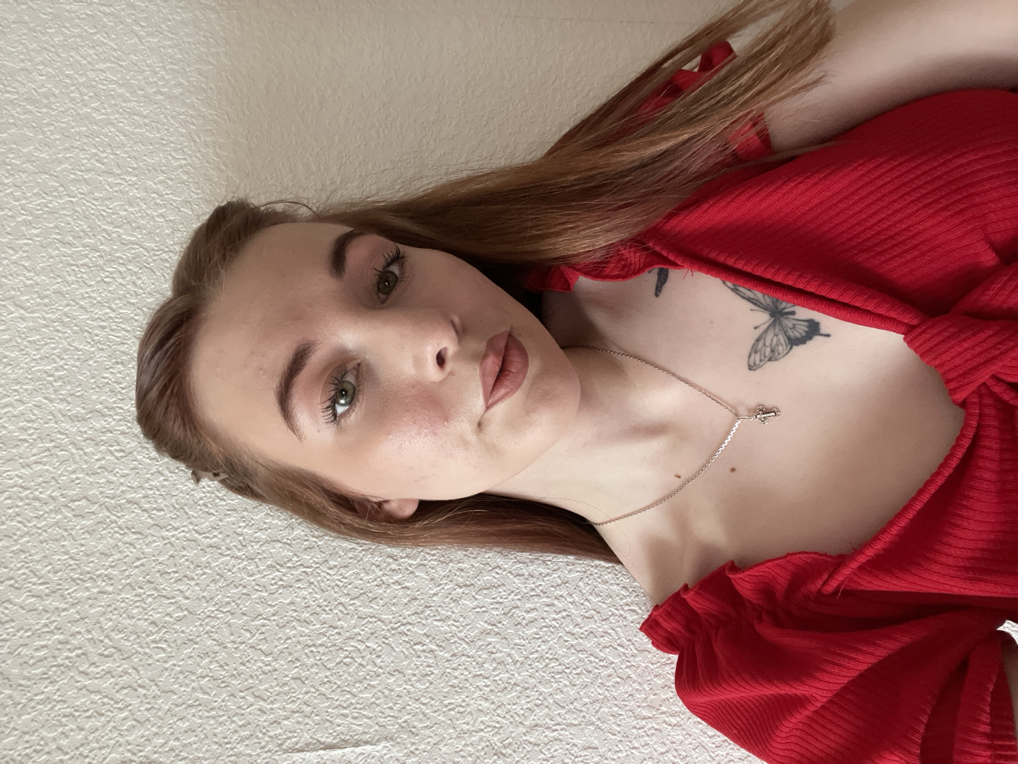 A fair-skinned woman with auburn hair and green eyes is photographed from a low angle. She is wearing a red, off-the-shoulder top, and a silver necklace with a small cross pendant. A butterfly tattoo is visible on her upper chest. The background is a white, textured wall. The location is Unknown, Unknown.