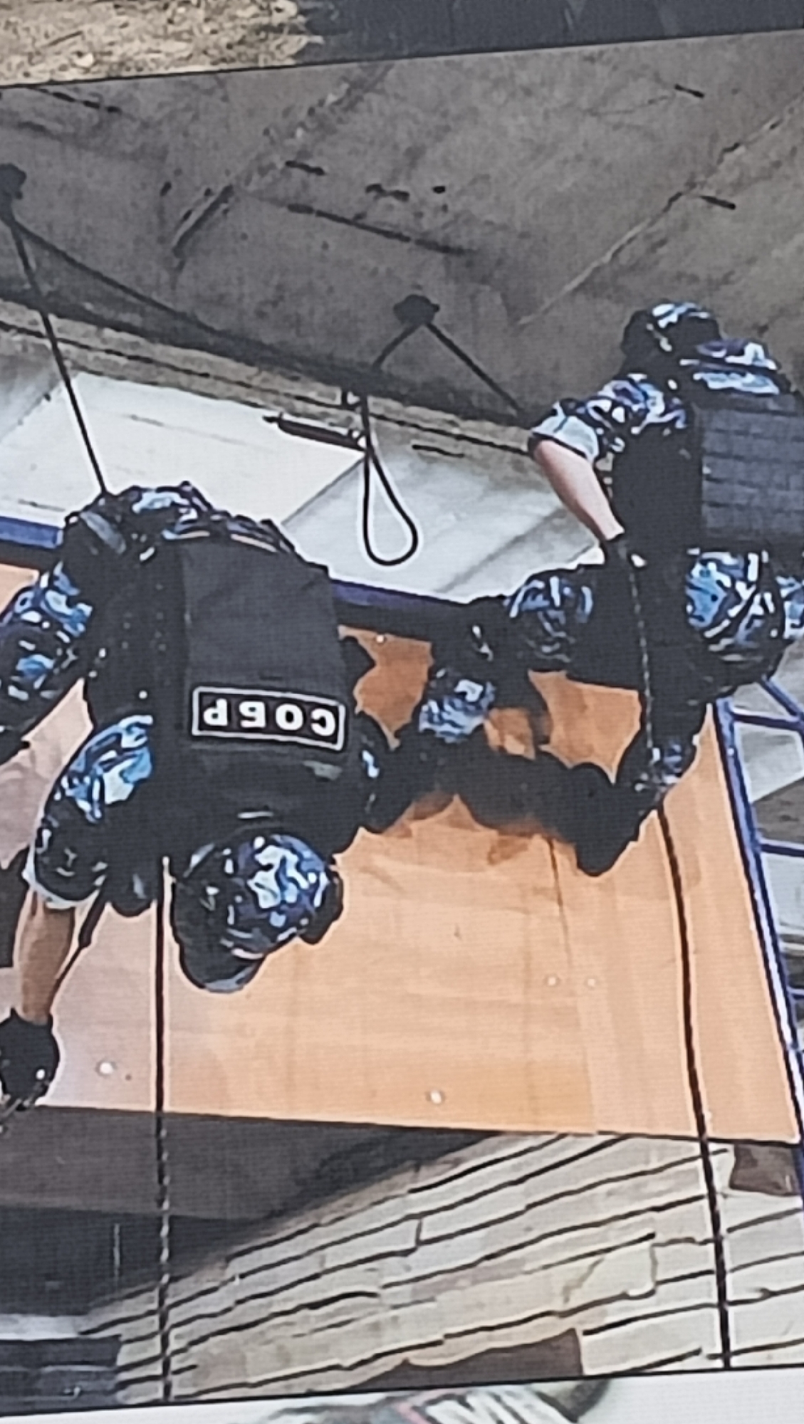 The image depicts two individuals, likely special forces or law enforcement personnel, engaged in a rappelling or fast-roping exercise. The photo is taken from below, showing the subjects as if suspended upside down against a ceiling, suggesting they are either practicing a head-first "Australian rappel" or the image is rotated to show them from a ground perspective.
Both individuals are equipped in tactical gear, including camouflage uniforms with a distinctive blue, grey, and black pattern, tactical vests or plate carriers, and helmets. The individual on the left clearly displays a white patch on their tactical vest with black Cyrillic text, which reads "СОБР" (SOBR). This acronym stands for Special Rapid Response Unit, typically associated with Russian law enforcement special forces. This person is seen holding a rope with black tactical gloves. The second individual on the right is similarly outfitted, with a portion of a blue ladder visible near their legs.
The setting appears to be an indoor or covered training facility, characterized by a concrete ceiling from which the ropes are suspended. Below the rappelling personnel, there is a large, light-brown wooden-like panel or wall, and further down, a textured, possibly layered, surface. The scene conveys a rigorous training environment focused on specialized tactical skills. Given the location context of Tiraspol, Moldova, and the "SOBR" insignia, these individuals are likely members of a Transnistrian special police or military unit, reflecting the region's strong ties and similar organizational structures to Russia.