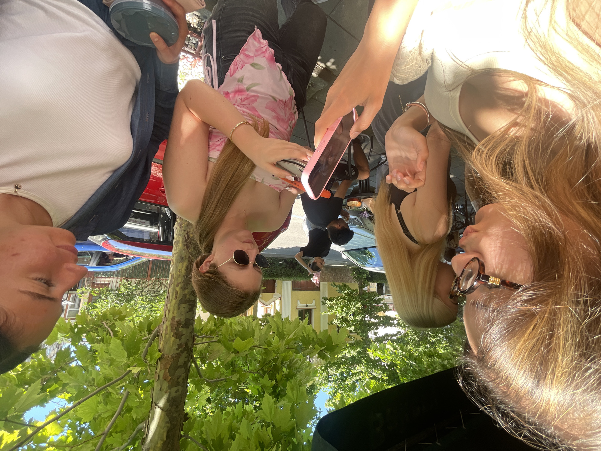 The image captures a street scene in Burgas, Bulgaria, on a sunny day. In the foreground, three young women are gathered. The woman on the far left, with long blonde hair, is wearing glasses and a light-colored top. Next to her, another woman with blonde hair is visible from the chest up, wearing a black top. The central figure, a woman with long blonde hair pulled back, is wearing a pink floral strapless top and black jeans. She is holding a pink phone in her left hand and a silver can in her right hand. To the right of her, another woman with dark hair is wearing a white ribbed top and a dark jacket. She is holding a dark oval container. In the background, trees with green foliage overhang the scene, and buildings with yellow and white facades are visible. Several cars, including a red one and a silver one, are parked on the street. A man in a black shirt and shorts is seated at a table in the background, with food on it. The overall atmosphere suggests a casual outdoor gathering.