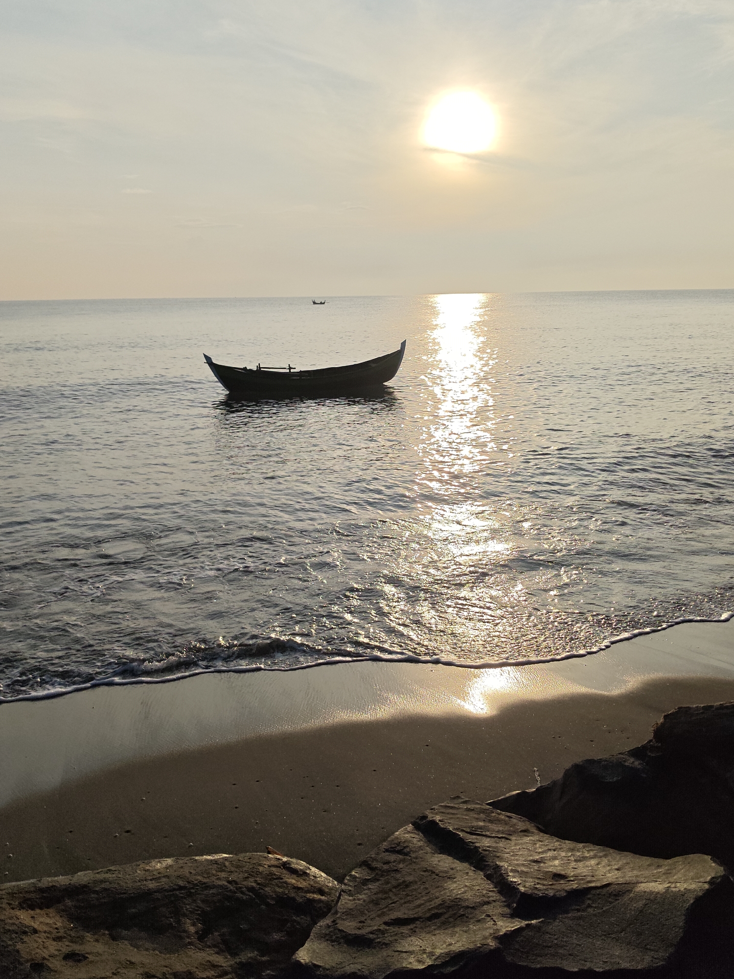 A long, narrow boat with a pointed bow floats on calm water. The sun is low in the sky, casting a bright reflection on the water. A sandy beach is in the foreground, with a few rocks. The scene is peaceful and serene. This image is likely taken at dawn or dusk at the Ujong Blang Beach in Lhokseumawe, Indonesia, a popular spot for watching the sunrise or sunset.