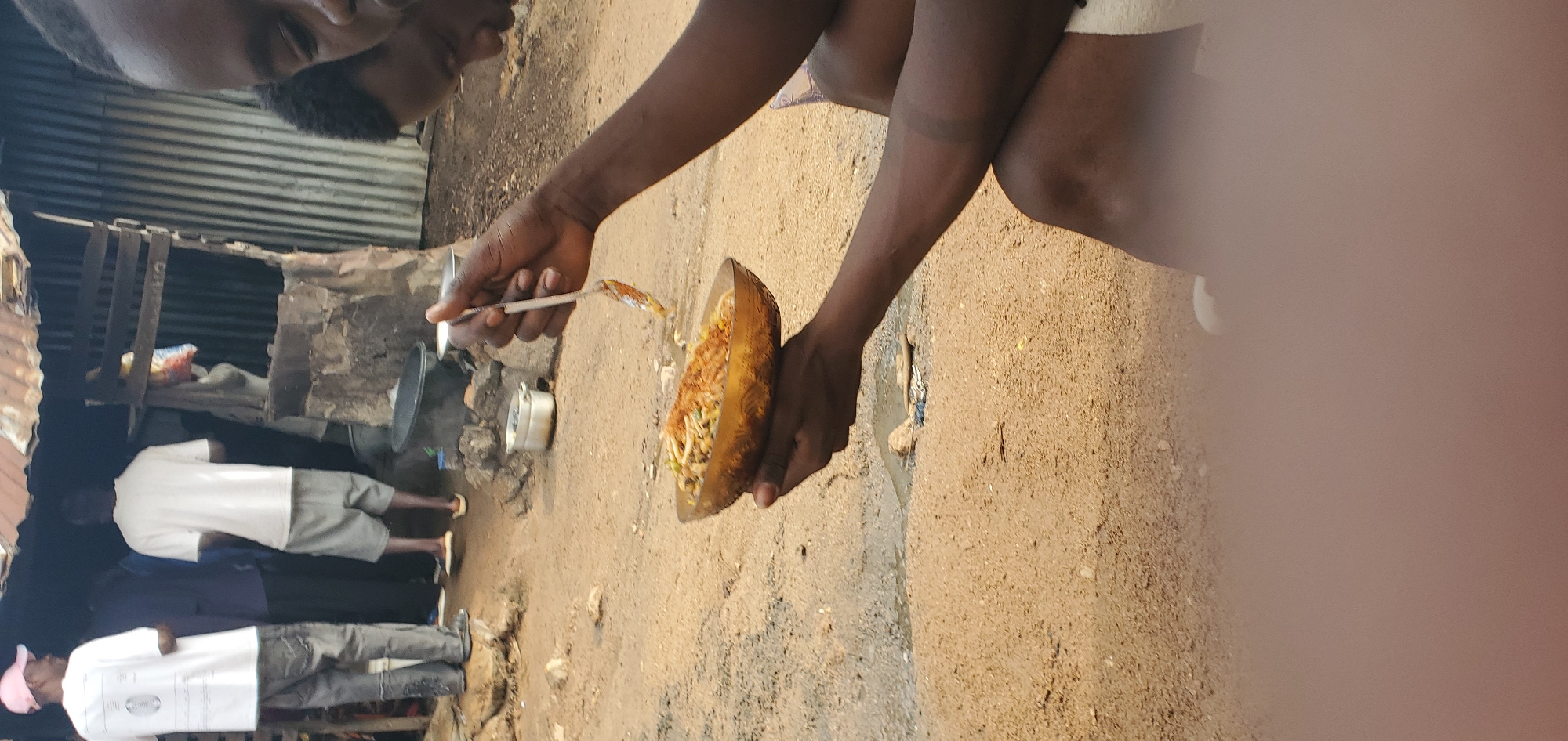 In an outdoor setting with a sandy ground, a person is holding a bowl of food with a spoon. The bowl is made of a golden-brown material and appears to contain noodles mixed with vegetables and spices. The person's hands are dark-skinned, and they are holding the bowl and spoon in a way that suggests they are about to eat or are in the process of serving themselves.
In the background, there is a rustic structure that appears to be a makeshift shop or food stall. It has a corrugated metal roof and walls made of wood and possibly stone. Several other people are visible in the background, some standing and some appearing to be interacting with the stall. One person is wearing a white t-shirt and a pink cap, another a white t-shirt and grey shorts. The environment looks like an informal urban or rural setting, possibly a market or a street food area. The overall impression is of a casual meal being prepared or consumed in a simple, outdoor environment.