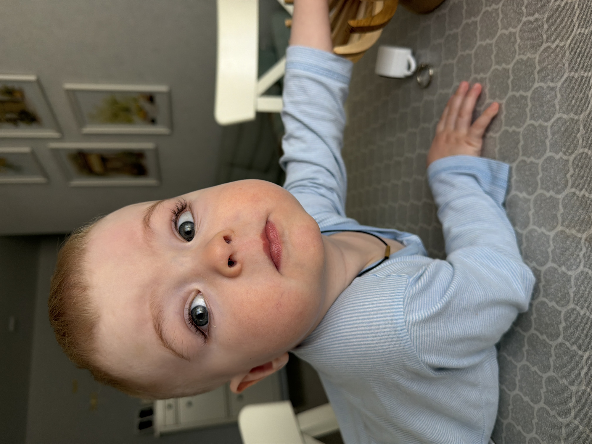 This close-up image, captured in Votkinsk, Russia, features a young child, likely a toddler, looking directly at the camera with a curious and engaging expression. The child has light reddish-blonde hair, fair skin, and large, expressive blue-grey eyes. Their lips are slightly parted, adding to their inquisitive gaze.
The child is wearing a light blue, long-sleeved shirt with subtle horizontal white stripes, and a thin dark cord necklace with a small golden bead is visible around their neck. One of their hands is resting on a grey surface covered with a repeating quatrefoil pattern, which appears to be a tablecloth or mat. Their other arm is extended upwards and to the left, towards a light-colored piece of furniture, possibly a chair or table leg, which is partially visible.
In the background, several framed pictures or artworks are mounted on a plain light-colored wall, though they are out of focus. A white ceramic mug and a silver key ring are visible on the patterned surface near the child's right hand. The indoor setting suggests a home environment. The perspective of the photograph gives the impression that the child is looking up from a low vantage point, perhaps lying on the floor or a low surface. The lighting is soft and even, suggesting either diffused daylight or artificial indoor lighting. No visible text is present in the image.