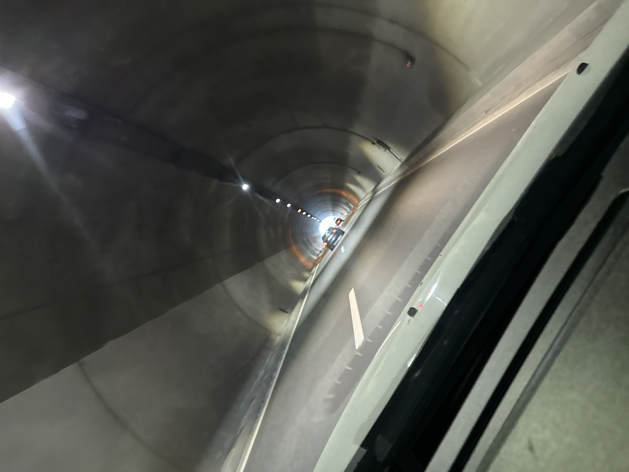 The image depicts the interior of a tunnel, likely a road tunnel, with a perspective from inside a vehicle. The tunnel walls are made of concrete and are illuminated by artificial lights, creating reflections on the road surface. There are lane markings visible on the road. Further down the tunnel, another vehicle can be seen, and at the far end, a brighter light source indicates the tunnel exit. The angle of the shot suggests a wide-angle lens or a fisheye effect, distorting the perspective. The scene is devoid of people other than implied occupants of the vehicles. The environment is sterile and functional, typical of modern tunnel infrastructure. The specific location in Blatino, Bulgaria, cannot be determined from the visual information alone, as there are no identifying landmarks or signs.