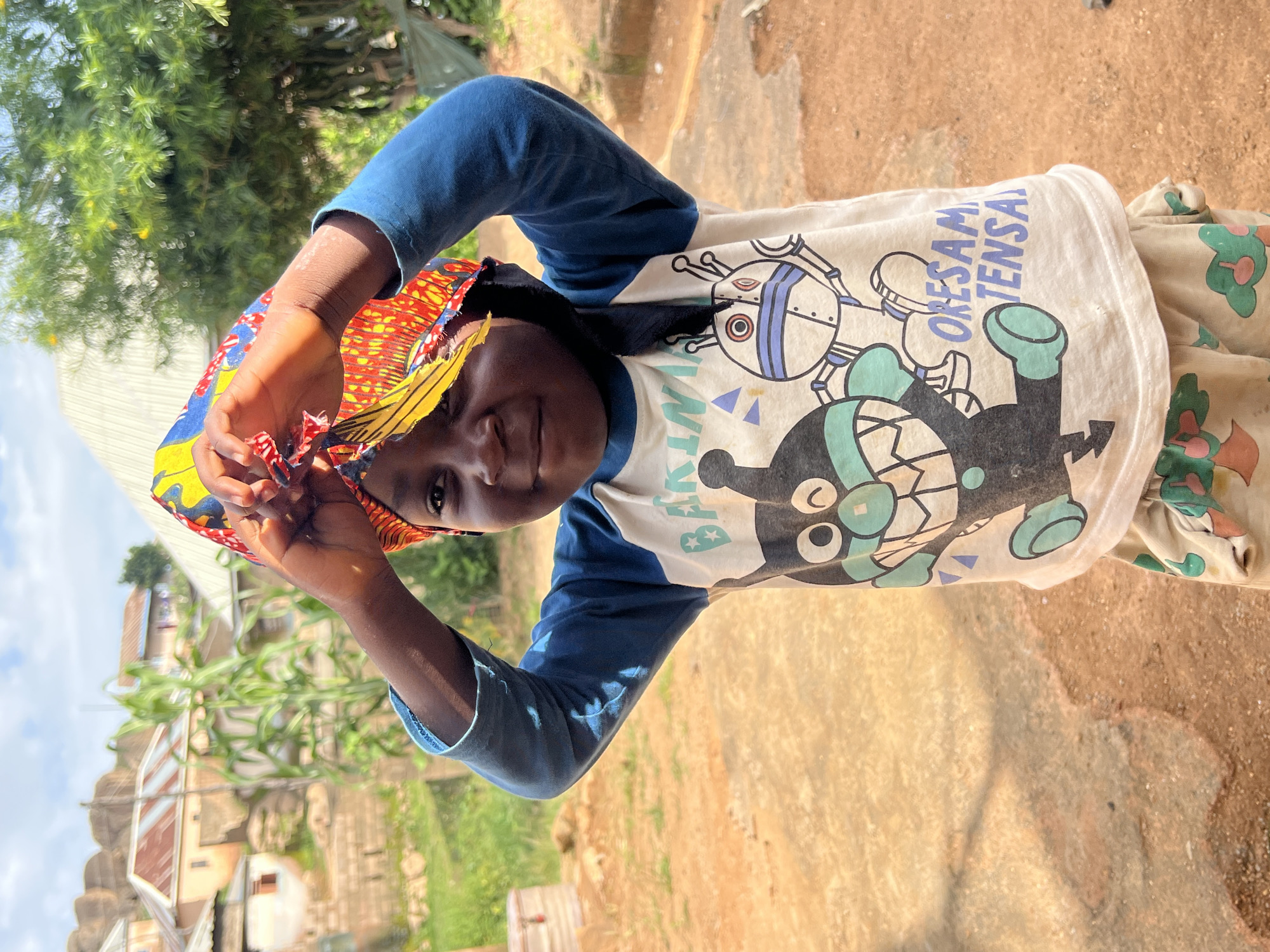 A young child, likely of African descent, stands outdoors, looking at the camera with a slight smile. The child is wearing a blue long-sleeved shirt over a white t-shirt adorned with cartoon characters and text. A colorful, patterned headwrap is positioned on the child's head, with the child's hands holding it in place. The background shows a village setting with buildings and vegetation, suggesting a rural or semi-urban environment. The ground is a reddish-brown dirt path. The lighting suggests it is daytime, with the sun casting some shadows. The scene appears to be located in Jos, Nigeria.