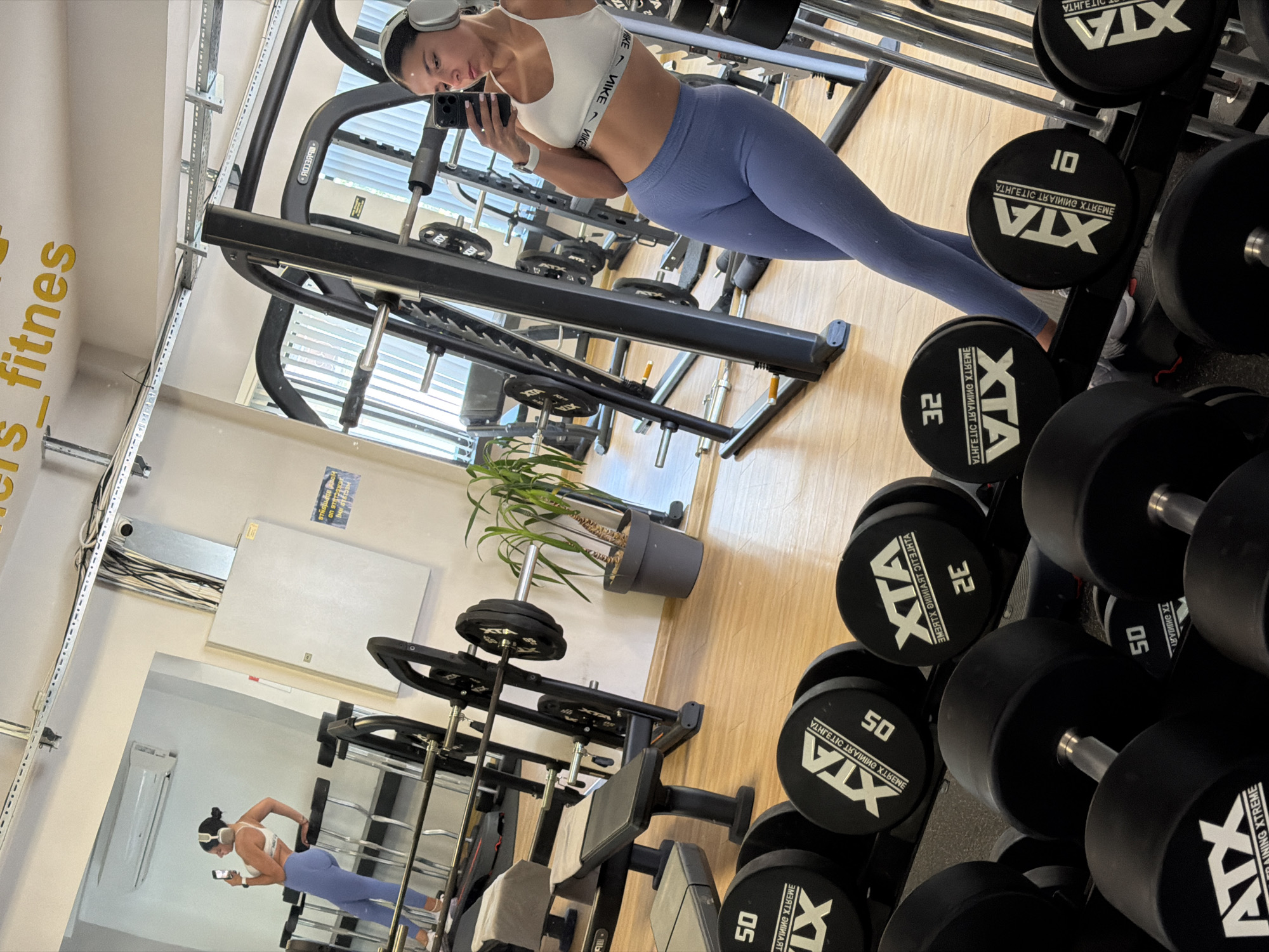 A young woman with dark hair tied up in a bun and wearing white headphones, a white sports bra, and light blue leggings stands in a gym, taking a mirror selfie. She is holding a smartphone in her right hand, capturing her reflection. The gym features wooden floors, weightlifting equipment including barbells and a rack, and various dumbbells. In the background, a plant in a dark pot is visible. The gym's ceiling has exposed electrical conduits and lighting fixtures. A sign with yellow lettering is visible on the wall. The overall environment suggests a fitness facility. The location is identified as "Fitnes Masters" in Gabrovo, Bulgaria.