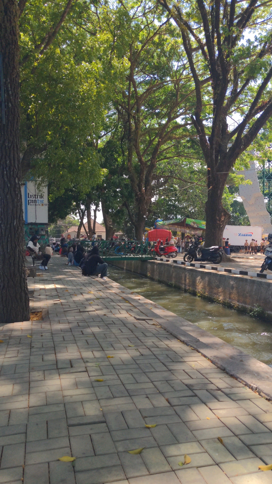 The image depicts a sunny day in a public area alongside a canal or small river in Magelang, Indonesia. A paved walkway runs along the canal, bordered by a low wall. Large trees with dense foliage provide shade over the walkway and surrounding area.
Several people are visible, primarily sitting or standing along the walkway. In the foreground, on the left, a large tree trunk is prominently featured. Further down the walkway, a group of individuals are seated, some facing the canal, others conversing. A sign for "Listrik Pintar" (Smart Electricity) is affixed to a structure on the left.
Across the canal, a green railing separates the walkway from parked motorcycles, several of which are red scooters. In the background, there are stalls or temporary structures, indicating a market or event. A white truck with the logo "Ziano" is parked on the far side, and behind it, a large white climbing wall structure is visible. The canal water appears to be flowing. The overall scene suggests a recreational or commercial area frequented by locals.