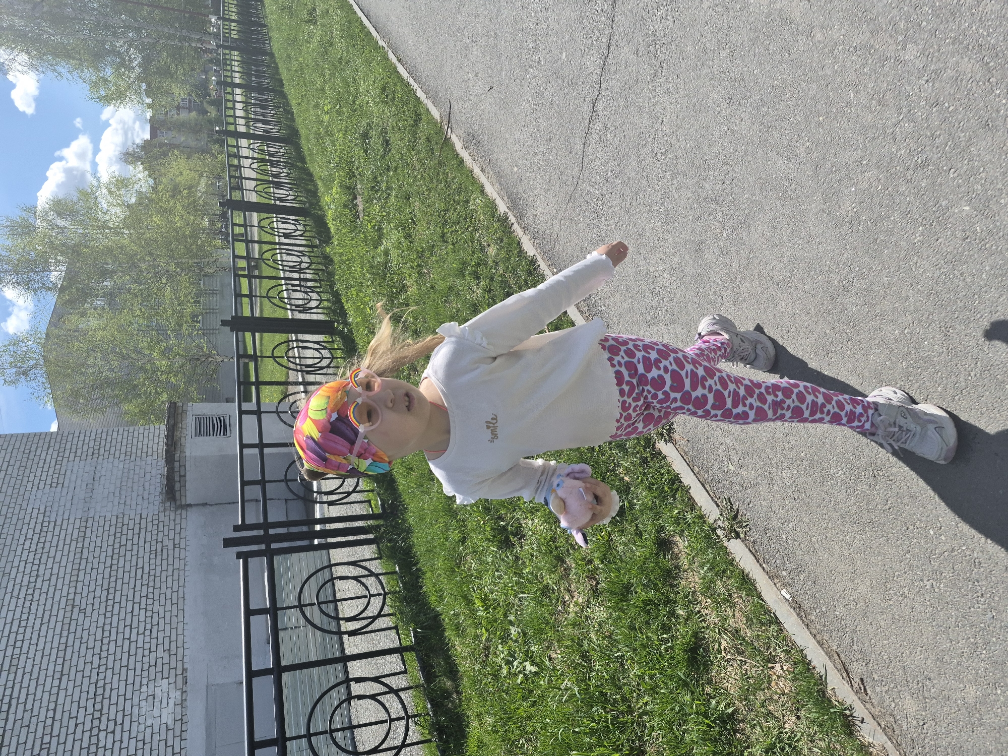 A young girl stands on a sidewalk adjacent to a grassy area bordered by a black metal fence. The sidewalk runs parallel to the fence and then a brick building. She wears a white, long-sleeved shirt, pink leggings with white heart prints, and gray sneakers. A multicolored headscarf covers her hair, and she wears rainbow-rimmed sunglasses. She holds a small, pink, plush object. The setting includes a paved sidewalk, a strip of green grass, a dark fence, and a brick building under a sky with scattered clouds. Based on the location, the image is likely from Tobolsk, Russia.