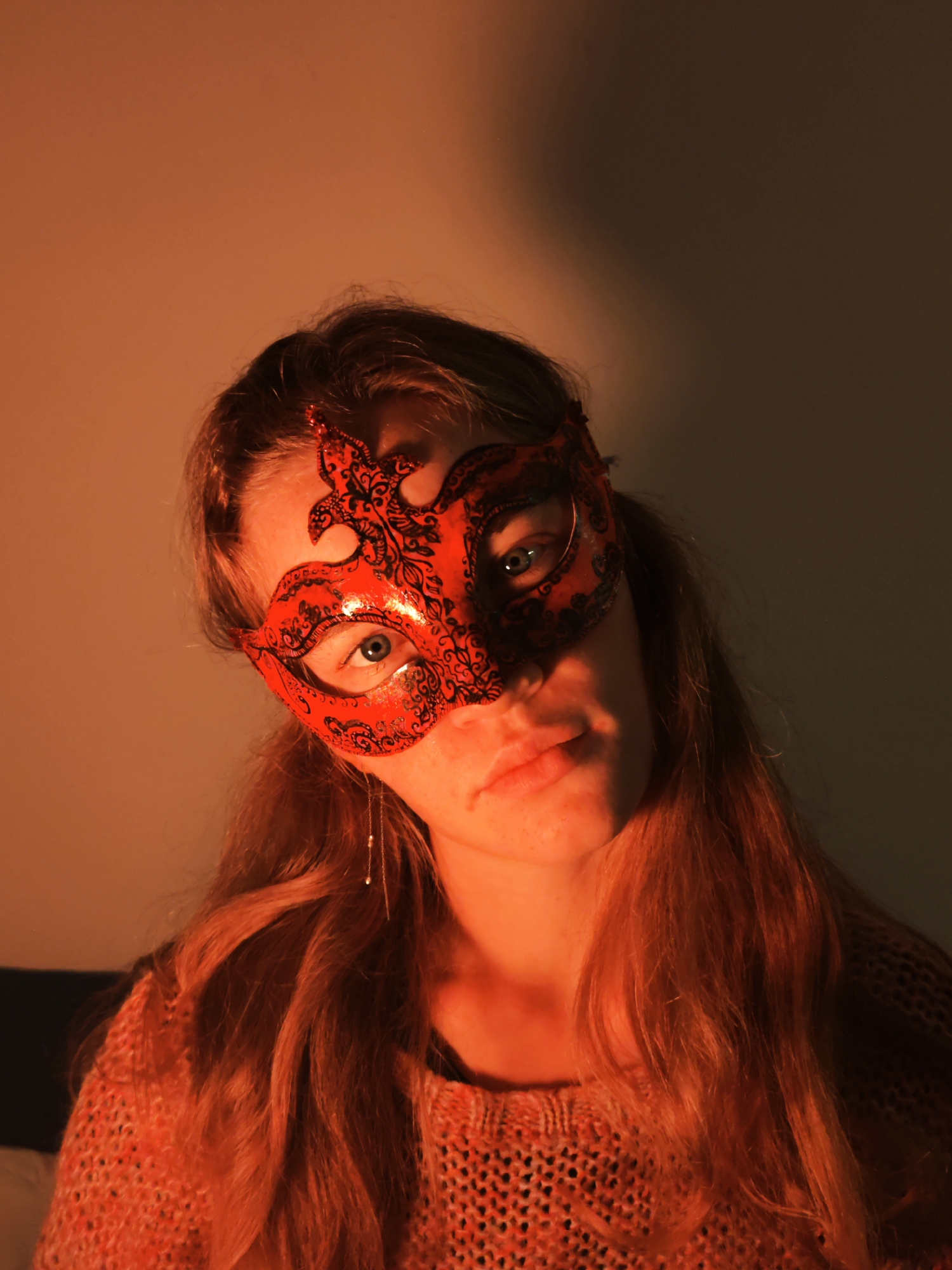 A close-up, waist-up portrait shows a light-skinned woman wearing a red mask with black embellishments. The mask covers the upper half of her face, revealing blue eyes. Her long, reddish-brown hair is loose. She is wearing a knitted sweater in a similar reddish hue. The lighting is warm, creating a slightly orange tone across the entire image. The background is a plain wall, with a dark object visible in the lower left corner. No specific location or indications of Budva, Montenegro, are discernible.