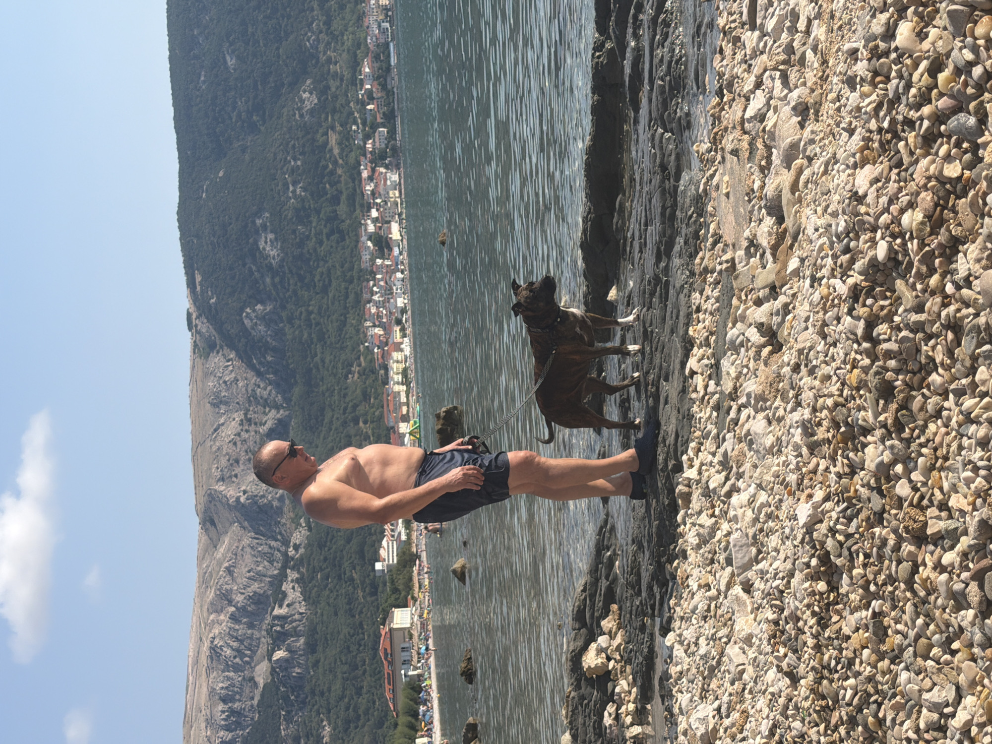 An outdoor, full shot captures a man and his dog on a rocky shoreline in Baška, Općina Baška, Croatia. The man, bare-chested and wearing dark blue swim trunks and dark blue slip-on shoes, stands facing left with sunglasses on. He is holding a leash attached to a medium-sized brown and white dog standing to his right. The dog faces left, looking away from the camera. The shoreline is covered in small, multi-colored pebbles and larger, dark gray rocks. The water is clear and slightly rippled. In the background, a dense green forest covers the slopes of a mountain range. A town with red-roofed buildings is visible along the coast, with a beach lined with umbrellas and people further in the distance. The sky is clear blue with a few white clouds.