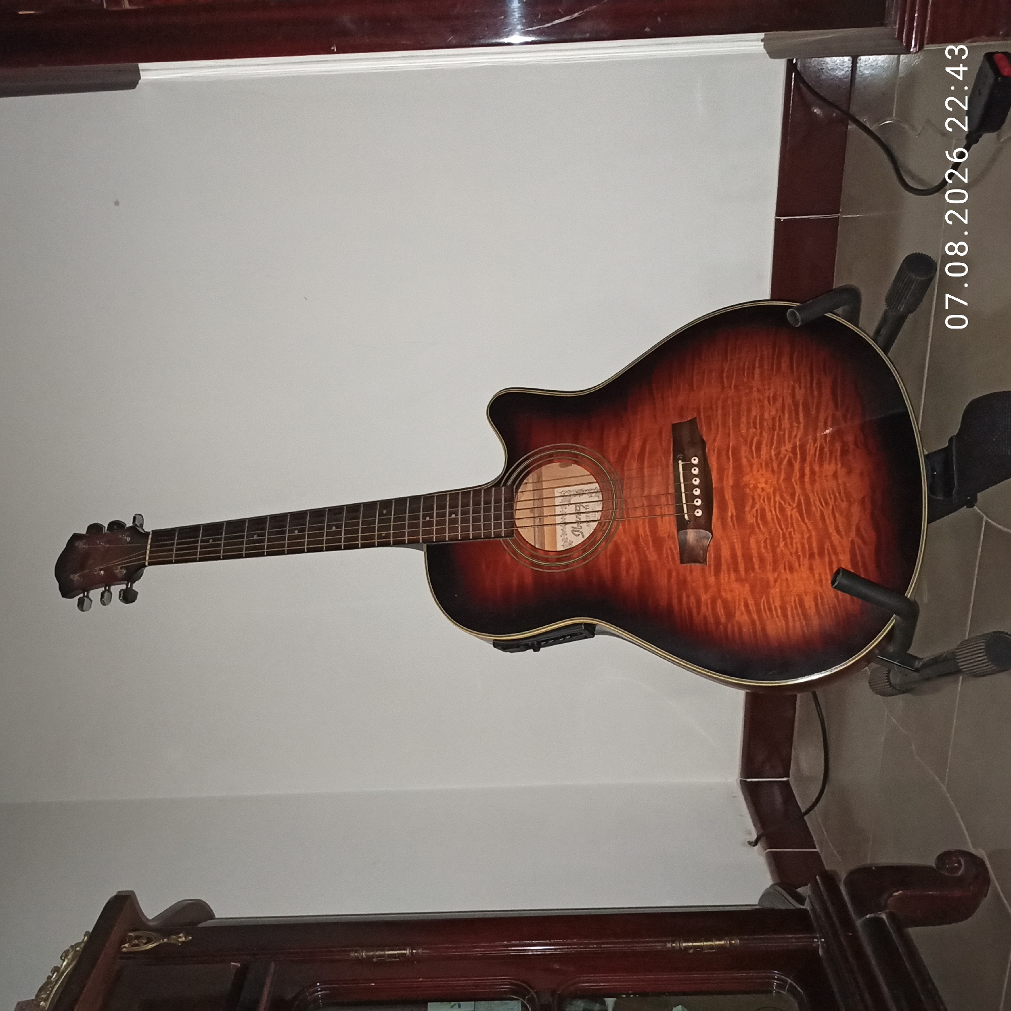 An acoustic-electric guitar with a sunburst finish and quilted maple top rests on a black guitar stand in a corner of a room. The guitar has a dark wood neck and fretboard, with six tuning pegs visible on the headstock. A black pickup is attached to the side of the guitar. To the left of the guitar is a tall, dark wooden cabinet with ornate gold trim. The wall behind the guitar is white, and the floor is tiled in a gray, marble-like pattern. A black cable runs from the guitar to a black power adapter on the floor. A timestamp in the lower right corner reads "07.08.2026 22:43". The scene appears to be indoors, possibly a home or studio, with no indication of the city of Bitonto, Italy.