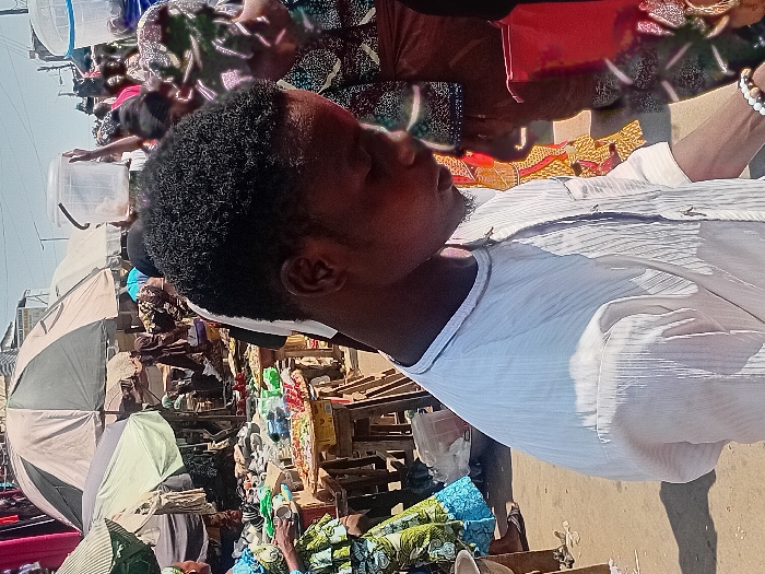 The image captures a vibrant, bustling outdoor market scene during bright daylight. In the foreground, a young Black man stands prominently, facing slightly to his right, with a calm or contemplative expression. He has short, dark, curly hair and a beard, and is wearing a light-colored, possibly white or pale blue, striped collared shirt. A bracelet with dark and light beads is visible on his right wrist.
Behind him, the market stretches out, filled with numerous stalls and people. Large, open umbrellas, including gray and white ones, provide shade over various vendor areas on the left side of the frame. The market is active with individuals, some of whom are blurred in the background, moving between stalls or examining merchandise. Various goods are displayed, including stacks of colorful patterned fabrics, plastic containers, and other assorted items arranged on tables and the ground. The ground appears to be unpaved dirt or sand. The clear, bright sky indicates a sunny day, casting strong natural light over the scene. The overall impression is that of a lively daily market in a populated area, characteristic of Magongo, Nigeria. No discernible text is visible within the image.