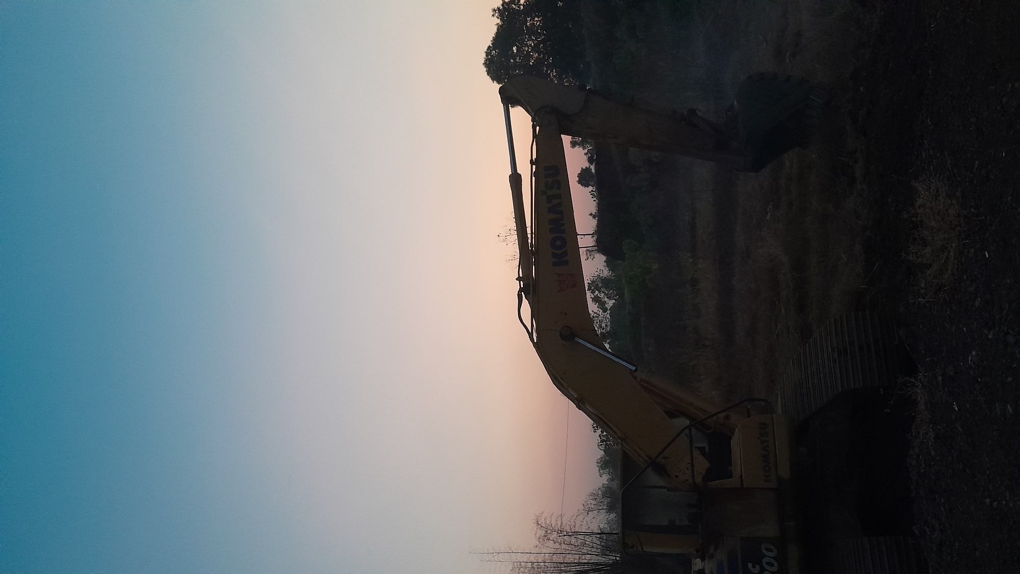 The image depicts an excavator parked in a field at dusk, likely in Sumberkramat, Indonesia. The sky is a gradient of blue and pale pink, transitioning to a lighter hue towards the horizon. The excavator, predominantly yellow, is positioned in the foreground, with its arm extended. The Komatsu logo is visible on the side of the arm. In the background, trees and other vegetation are silhouetted against the fading light. The ground is covered with dry grass and dirt, suggesting a rural or construction site environment. There are no people visible in the image. The overall atmosphere is serene and quiet, characteristic of twilight in a rural setting.
