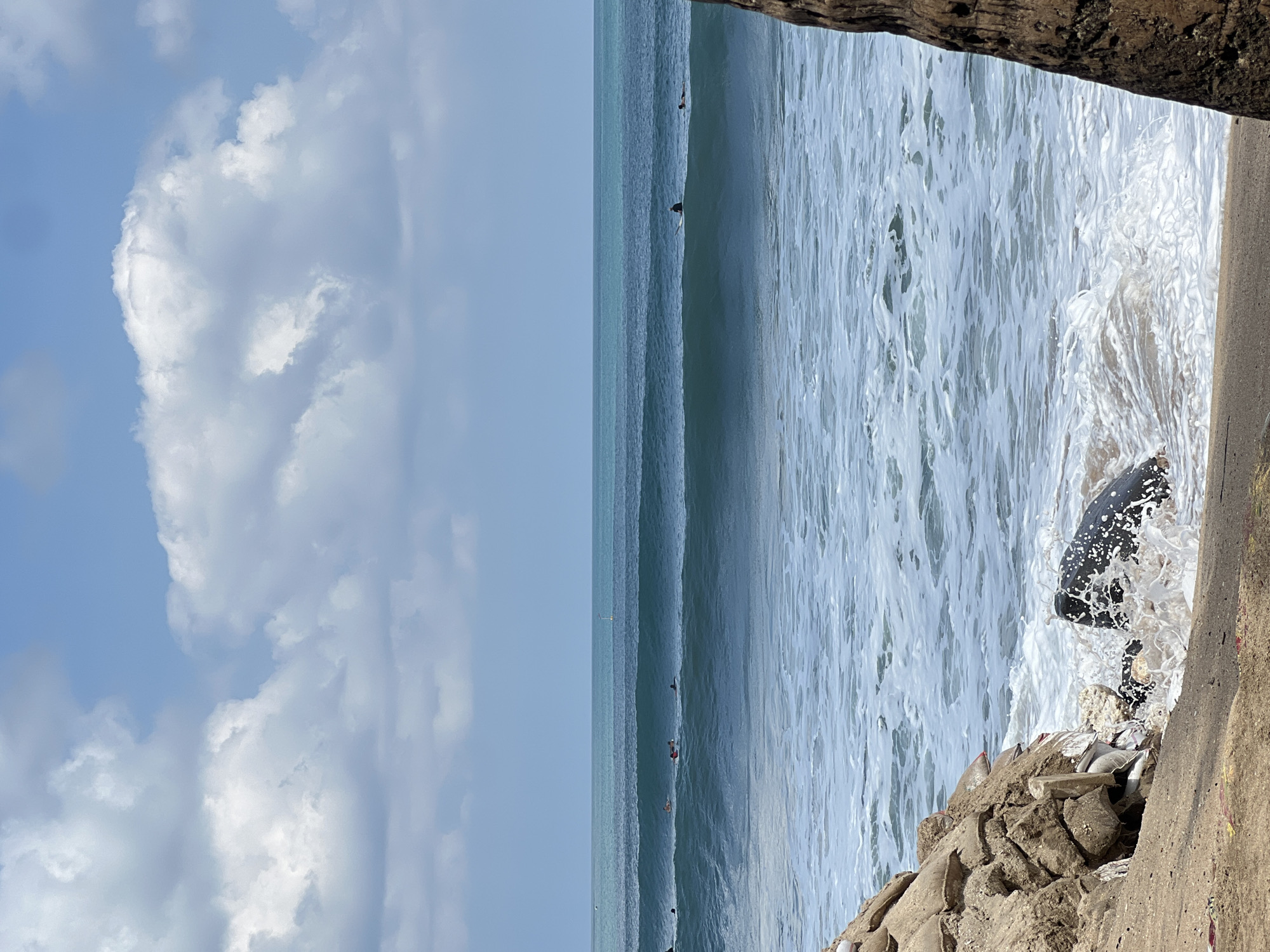 The image depicts a view of the ocean in Kuta, Indonesia, characterized by a bright, partly cloudy sky and calm, turquoise waters. Several surfers are scattered across the middle ground, patiently waiting for waves. The foreground of the image features a sandy beach littered with debris, including sandbags, plastic waste, and a large black object partially submerged in the crashing waves. A tree trunk is visible on the right side of the frame, adding a natural element to the scene. The overall atmosphere suggests a typical day at a popular surfing destination, though the presence of litter indicates environmental concerns.