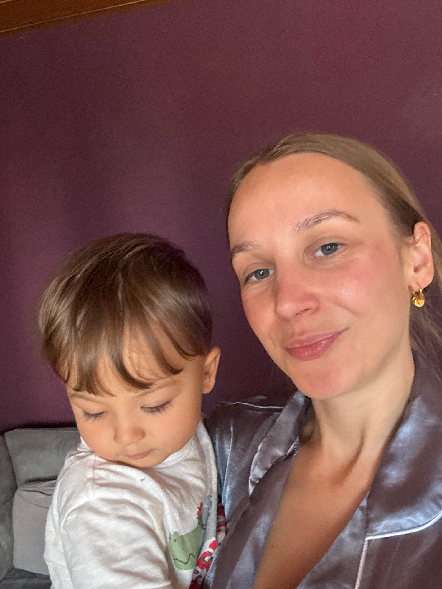 In a close-up, selfie-style photograph, a woman with blonde hair and blue eyes is holding a young child. The woman is wearing a grey satin pajama top with white trim. She has a gold hoop earring in her right ear. The child has brown hair and is looking down. The background consists of a dark purple wall and a portion of a gray sofa. The image appears to have been taken indoors, possibly in a residential setting in Überherrn, Germany.
