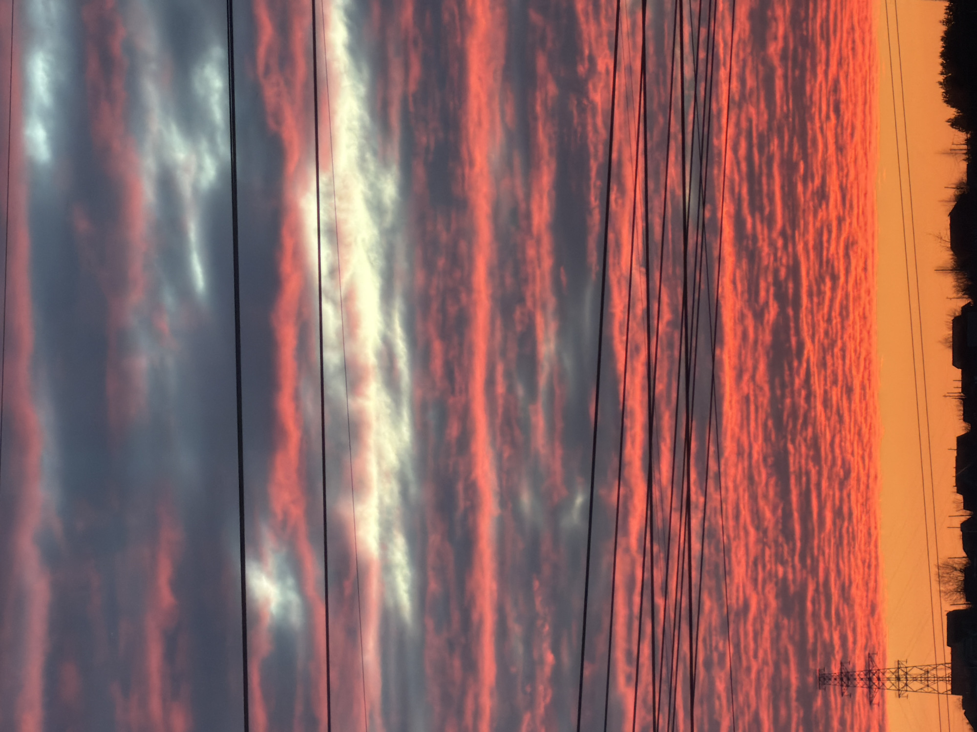 A wide-angle landscape photograph captures a sunset or sunrise over Chaikovsky, Russia. The sky is characterized by extensive horizontal cloud formations, predominantly altocumulus, displaying intense red and orange hues, with some upper clouds exhibiting grey and white illumination. Multiple dark, thin power lines traverse the image horizontally at various elevations. The lower portion of the scene presents a dark silhouette of the horizon, which includes indistinct residential or light commercial buildings, sparse leafless trees, and a prominent metal lattice transmission tower positioned on the left side. No individuals or active interactions are visible.