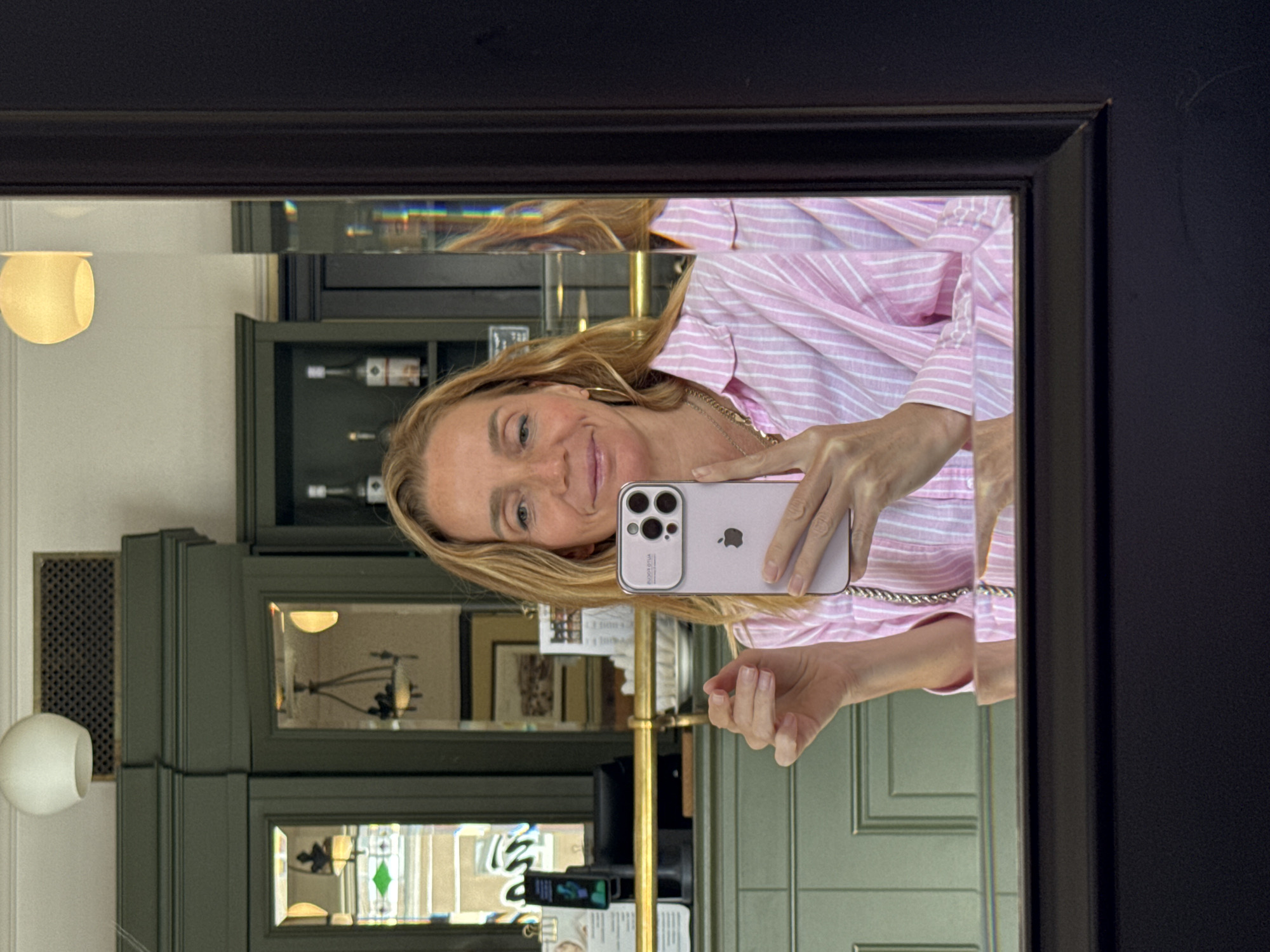 A woman is taking a mirror selfie in what appears to be a cafe or bar in Saint Petersburg, Russia. She is wearing a pink and white striped button-down shirt and gold hoop earrings. She holds a pink iPhone in her right hand. Reflected in the mirror behind her are dark green cabinets, shelves with bottles, and ornate light fixtures. The mirror itself is framed in black.