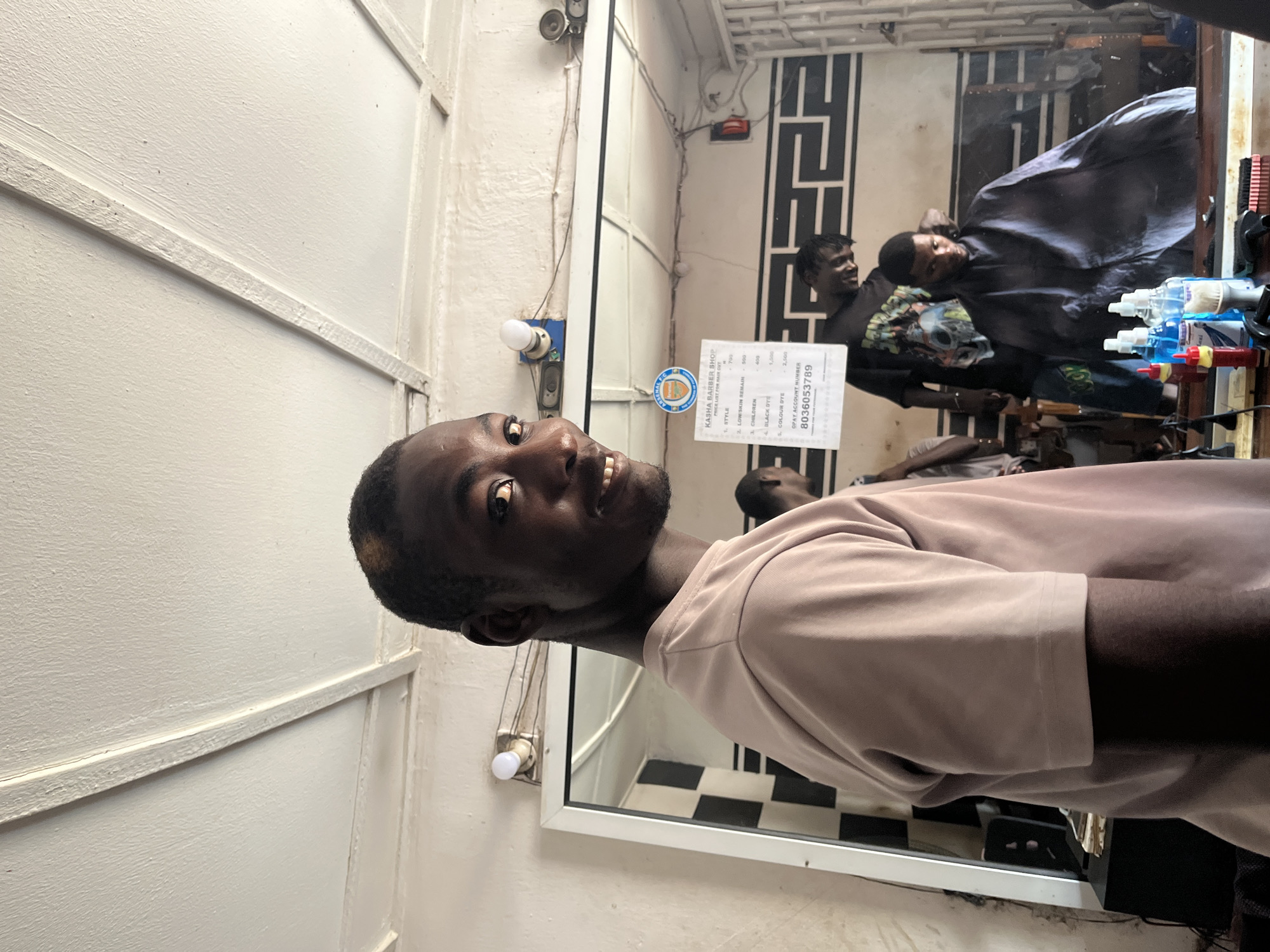 A person is standing in a barber shop in Jos, Nigeria. The shop has a mirror on the wall behind him. In the mirror, a barber is seen cutting another person's hair. There is a price list for hair cuts visible in the mirror as well with an OPAY account number of 8036053789. The floor has black and white tiles.