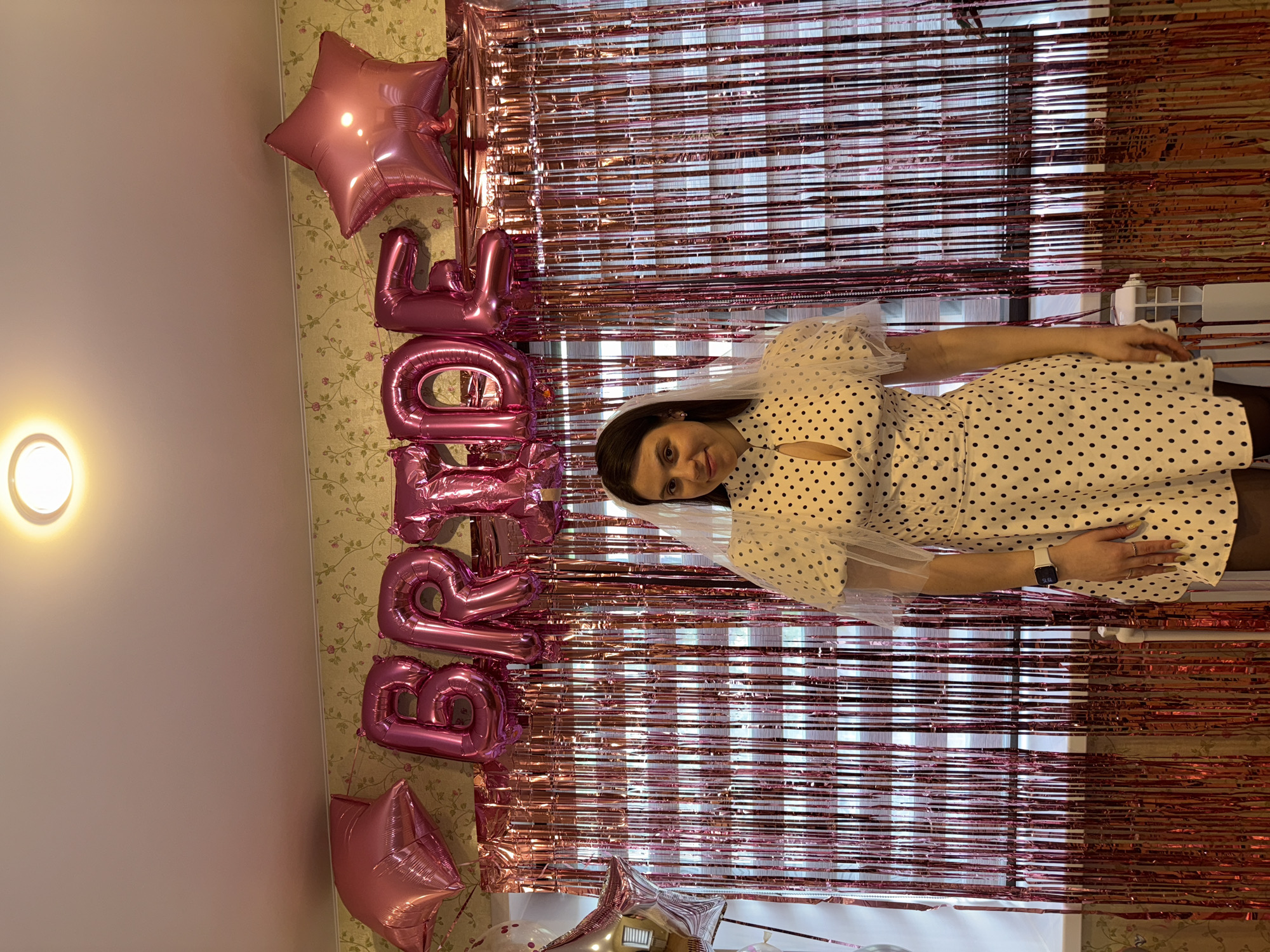 The scene depicts a young woman standing indoors, positioned centrally against a festive background. She wears a white dress adorned with black polka dots, featuring a high collar with a keyhole cutout and sheer white puff sleeves. A short white veil is draped over her dark hair. On her left wrist, she wears a white smartwatch, and rings are visible on her right hand. Her expression is a slight smile as she looks directly at the camera.
Behind her, a rose gold/pink tinsel curtain hangs, obscuring a window through which ambient light is visible. Above the tinsel curtain, strung across the wall, are pink foil balloons spelling out "BRIDE." These balloons are flanked by two large pink star-shaped balloons. To the far left, partially visible, are additional white and silver balloons, including another star shape. The wall above the balloons is covered with a light-colored wallpaper featuring a subtle floral pattern of green leaves and pink flowers on a greenish-yellow background. The ceiling, painted a plain light color, is visible at the top of the frame, featuring a circular flush-mount light fixture. Below the tinsel curtain, a white windowsill is visible, and beneath it, a segment of a white radiator unit. The setting is identified as being within the city of Perm, Russia.