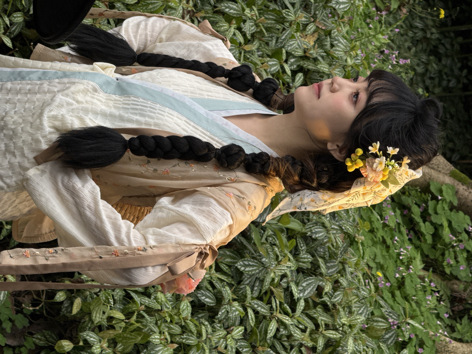 A woman lies on her side on a bed of mixed greenery in an outdoor setting. She has long, black hair styled into thick braids, adorned with small yellow and white flowers near her head. She wears a light-colored, textured garment with a light blue trim and an embroidered belt-like detail with ribbons. The foliage surrounding her includes plants with distinctive patterned leaves and small purple flowers. The location is the Yuzhong District, China.