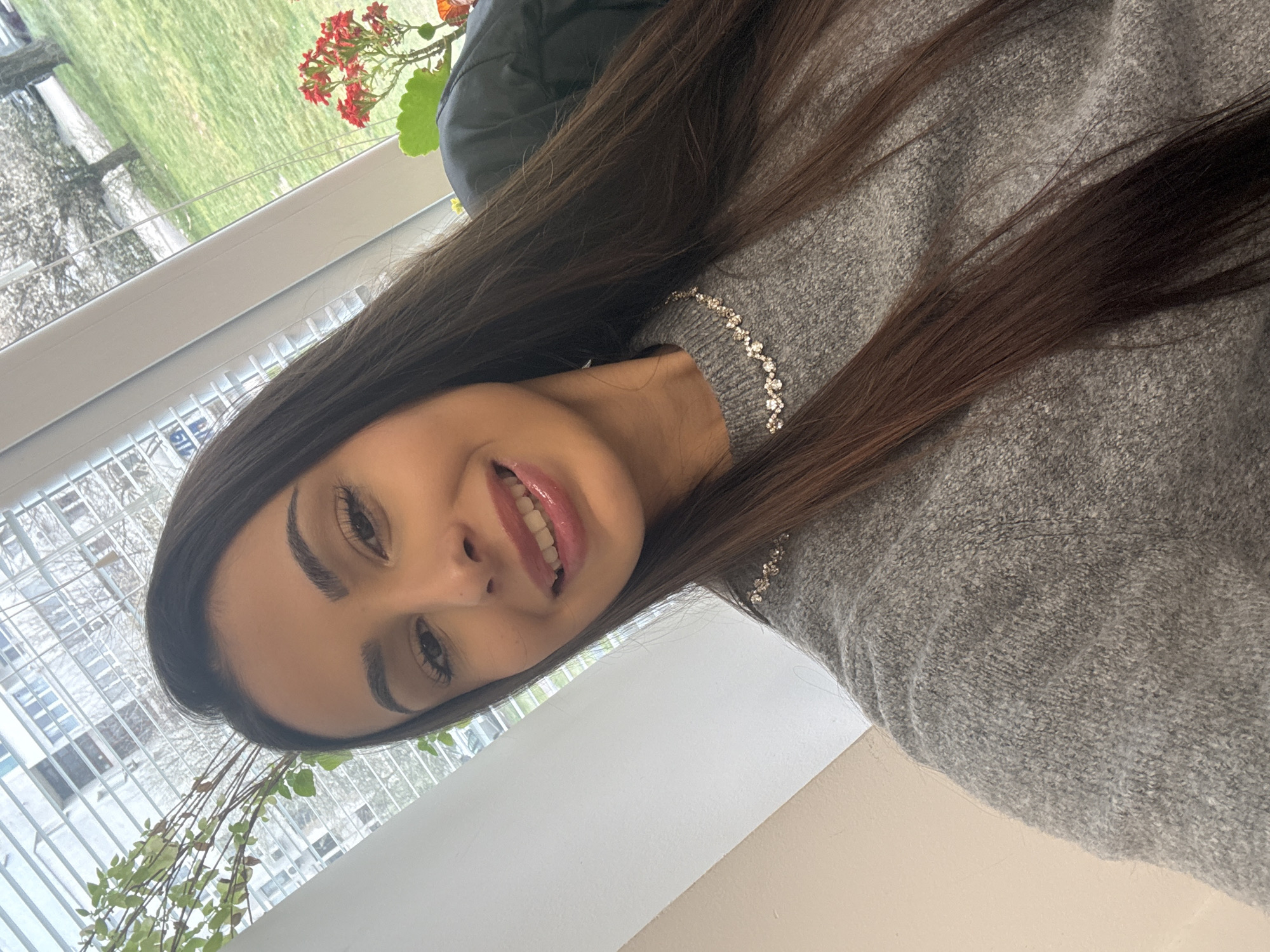 A woman with long, dark hair is taking a selfie. She is wearing a grey sweater with a sparkling neckline. The background shows a window with a view of greenery, possibly trees or plants. The location is indoors, near a window. There are no specific elements to identify the city of Sofia, Bulgaria.