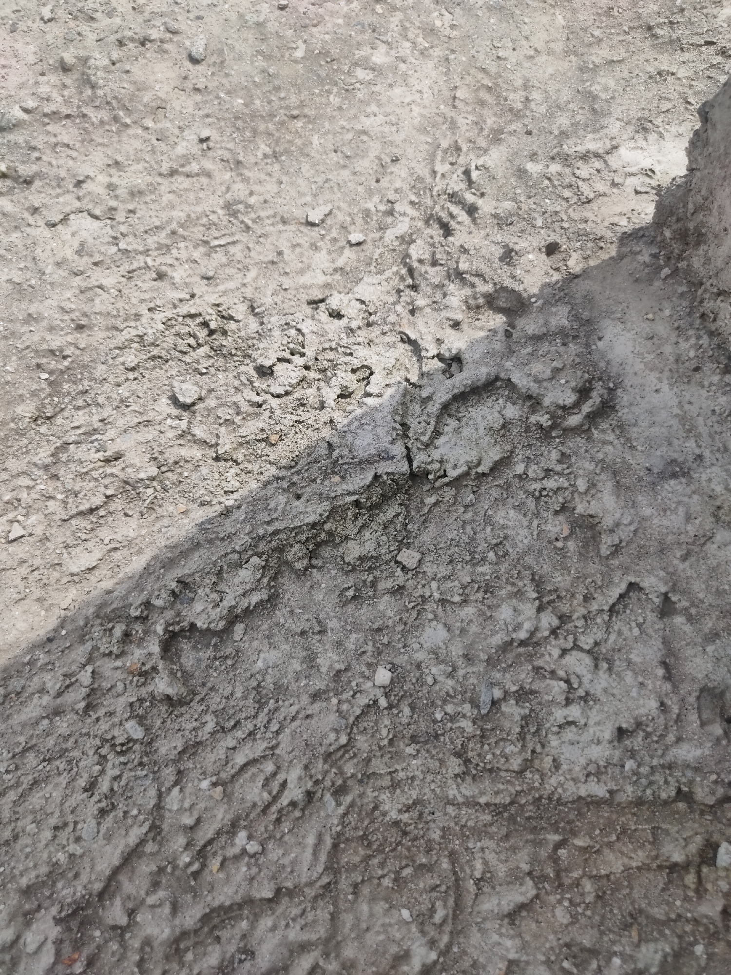 The image shows a close-up, eye-level view of a cracked and uneven concrete surface. The texture is rough, with small pebbles and irregularities visible. A shadow cuts diagonally across the surface, indicating a light source from the upper right. The concrete appears to be outdoors, possibly on a road or pathway. There are no people, animals, or other discernible objects in the frame. The overall impression is of a weathered and worn urban or semi-urban environment. The context of Manali, India, suggests this could be a section of pavement or an unpaved area in a town or on the outskirts, subject to the elements and possibly foot traffic.